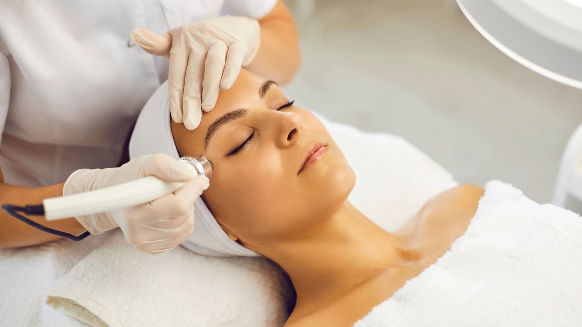 A woman receiving a facial treatment with a white facial device in a spa or aesthetic clinic. She is lying down with eyes closed, wearing a white headband and towel.