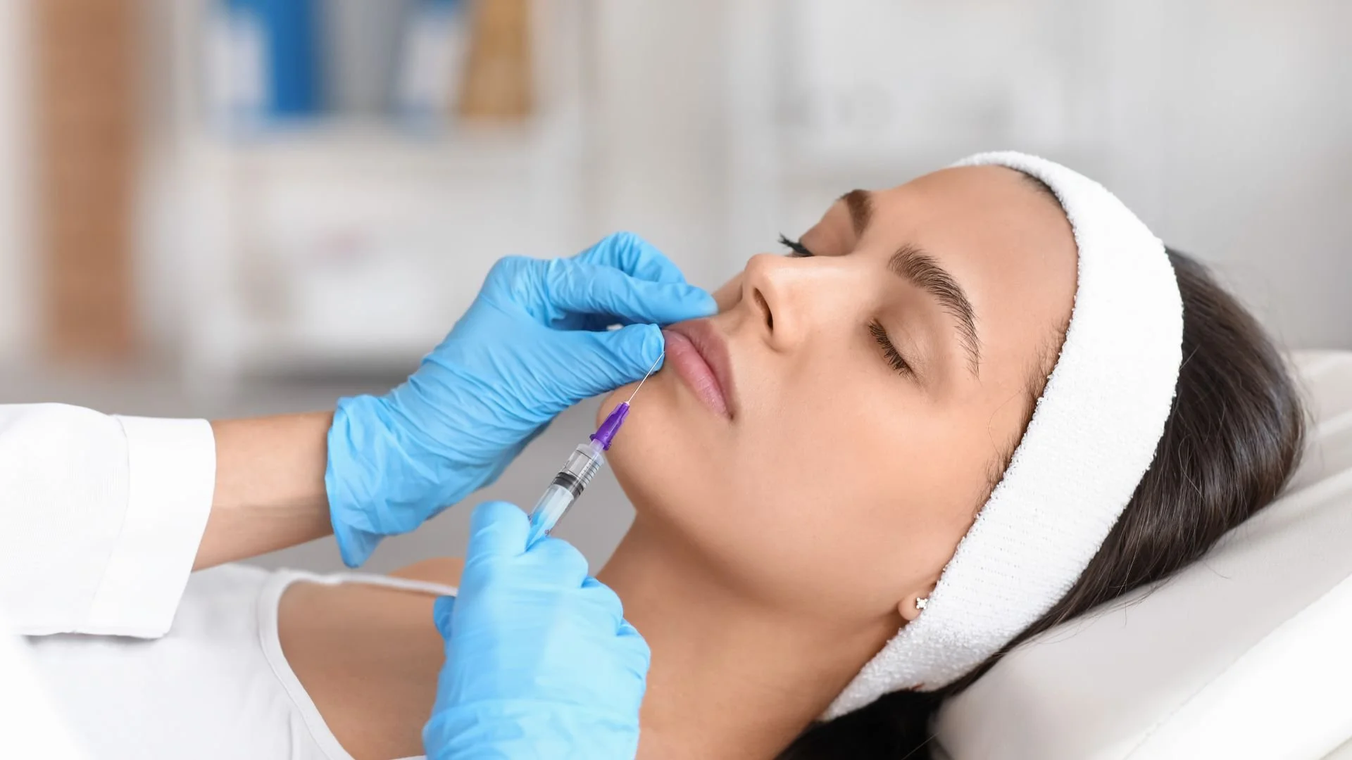 A woman receiving a cosmetic dermal filler facial treatment with a syringe administered by a professional wearing blue gloves, while lying down with a white headband and closed eyes.