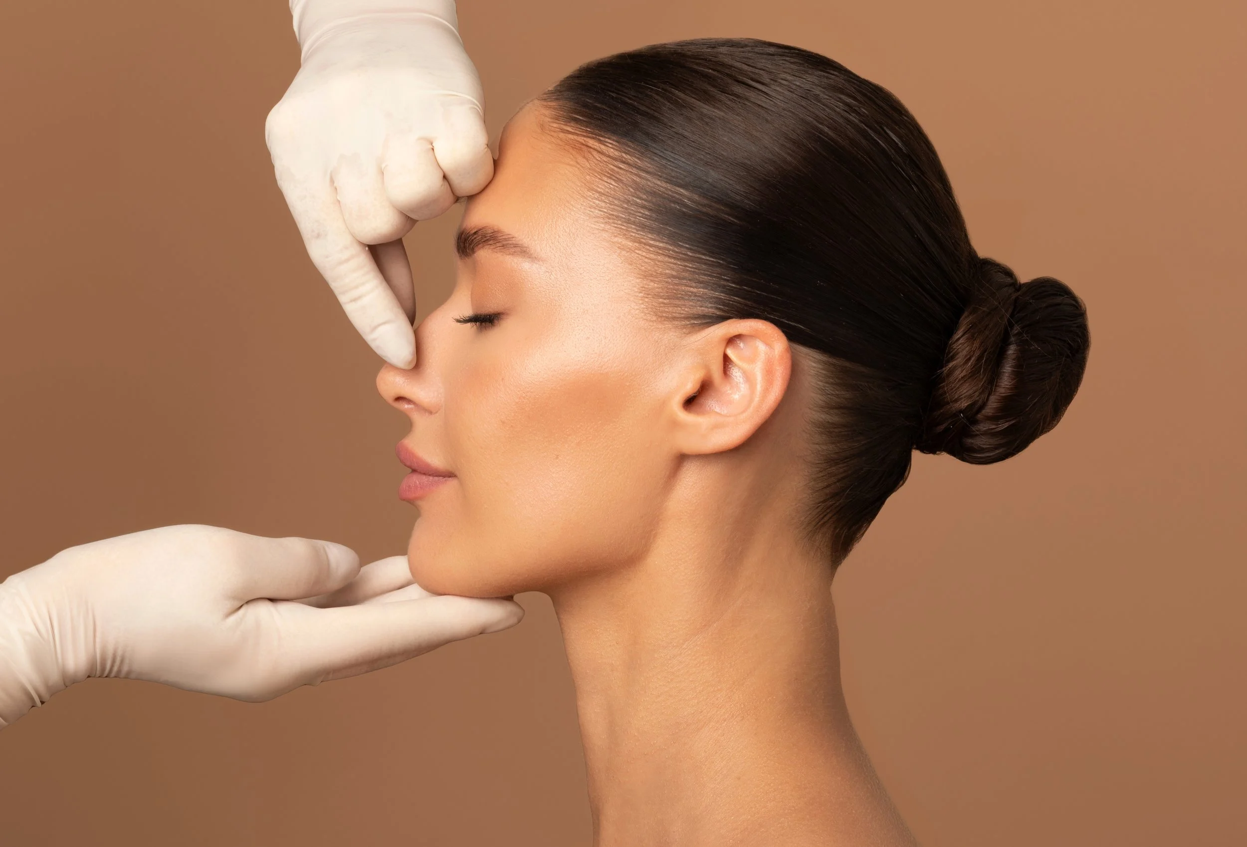 A woman with slicked-back hair and closed eyes has her nose and chin gently touched by gloved hands.