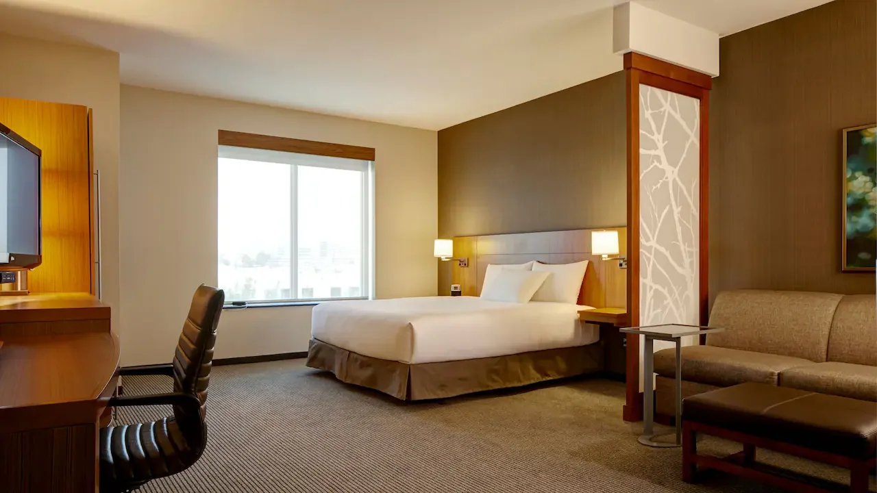 A modern hotel room featuring a large bed with white linens and a central brown pillow, bedside lamps, a beige chaise lounge, and a sofa with gray pillows, with neutral-colored walls and patterned carpet.