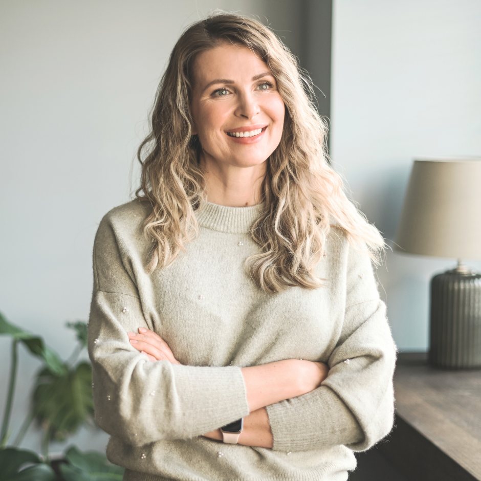 A woman with curly blonde hair smiling and crossing her arms in a cozy indoor setting.