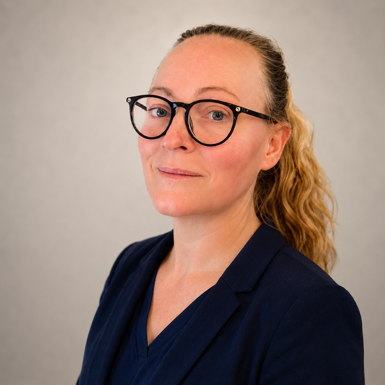 A professional woman with blond curly hair pulled back, wearing black-rimmed glasses and a navy blazer, looking confidently at the camera against a plain gray background.