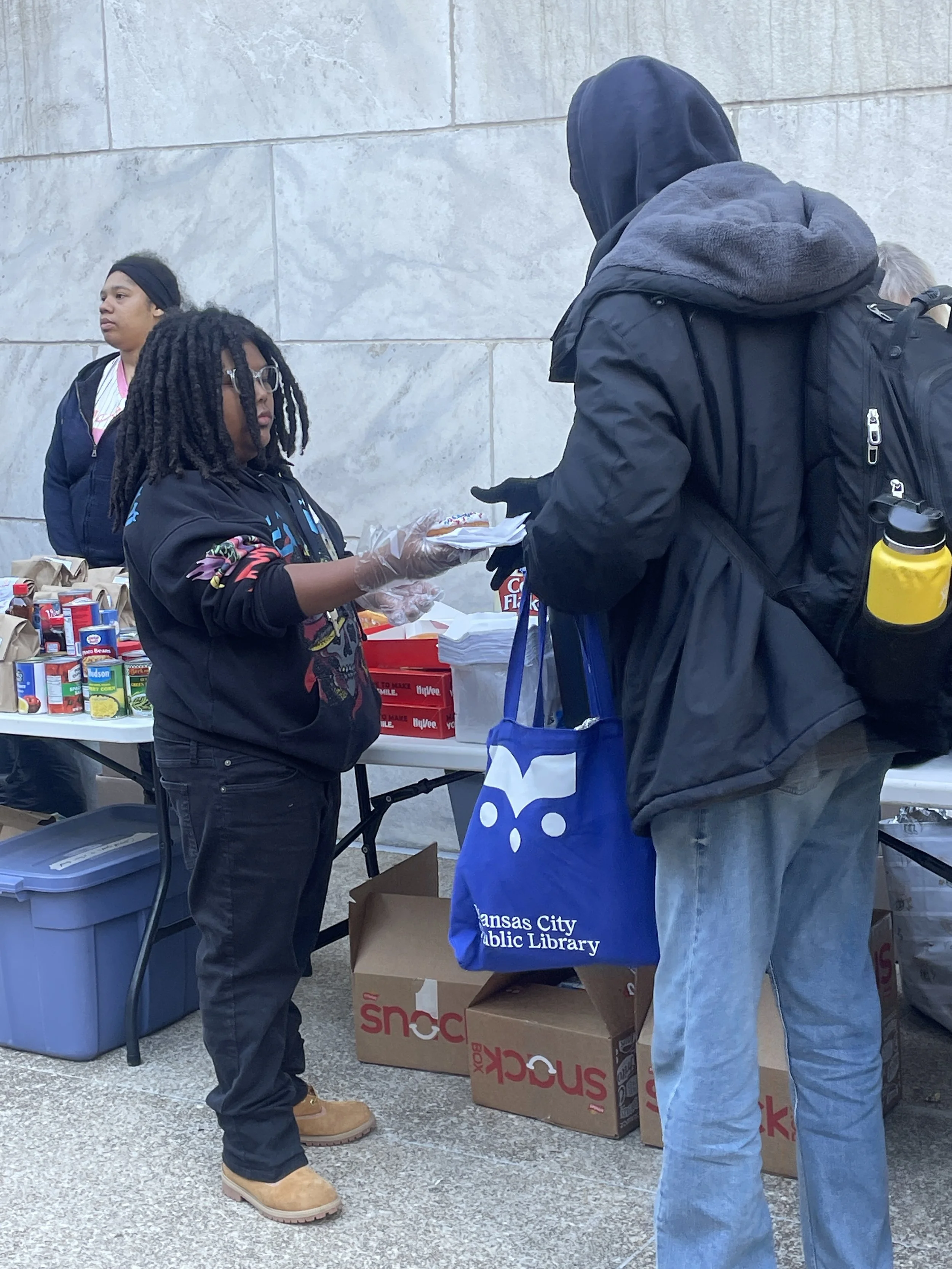 People at a food distribution event, with tables of canned goods and boxes, and one person handing something to another, in front of a gray stone wall.