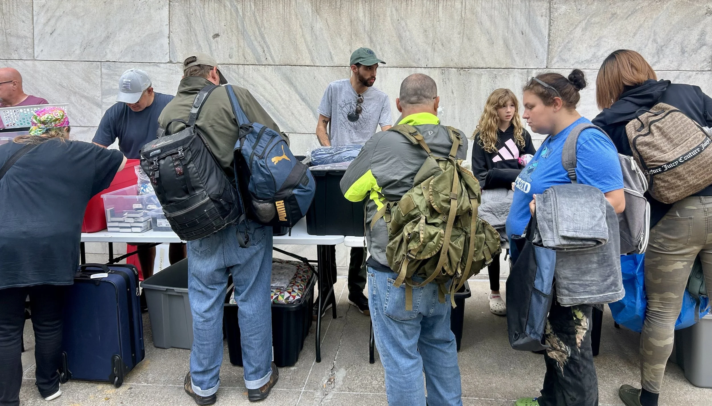 People gather around tables at a Street Support KC outreach event, with backpacks and luggage nearby, against a stone wall backdrop.
