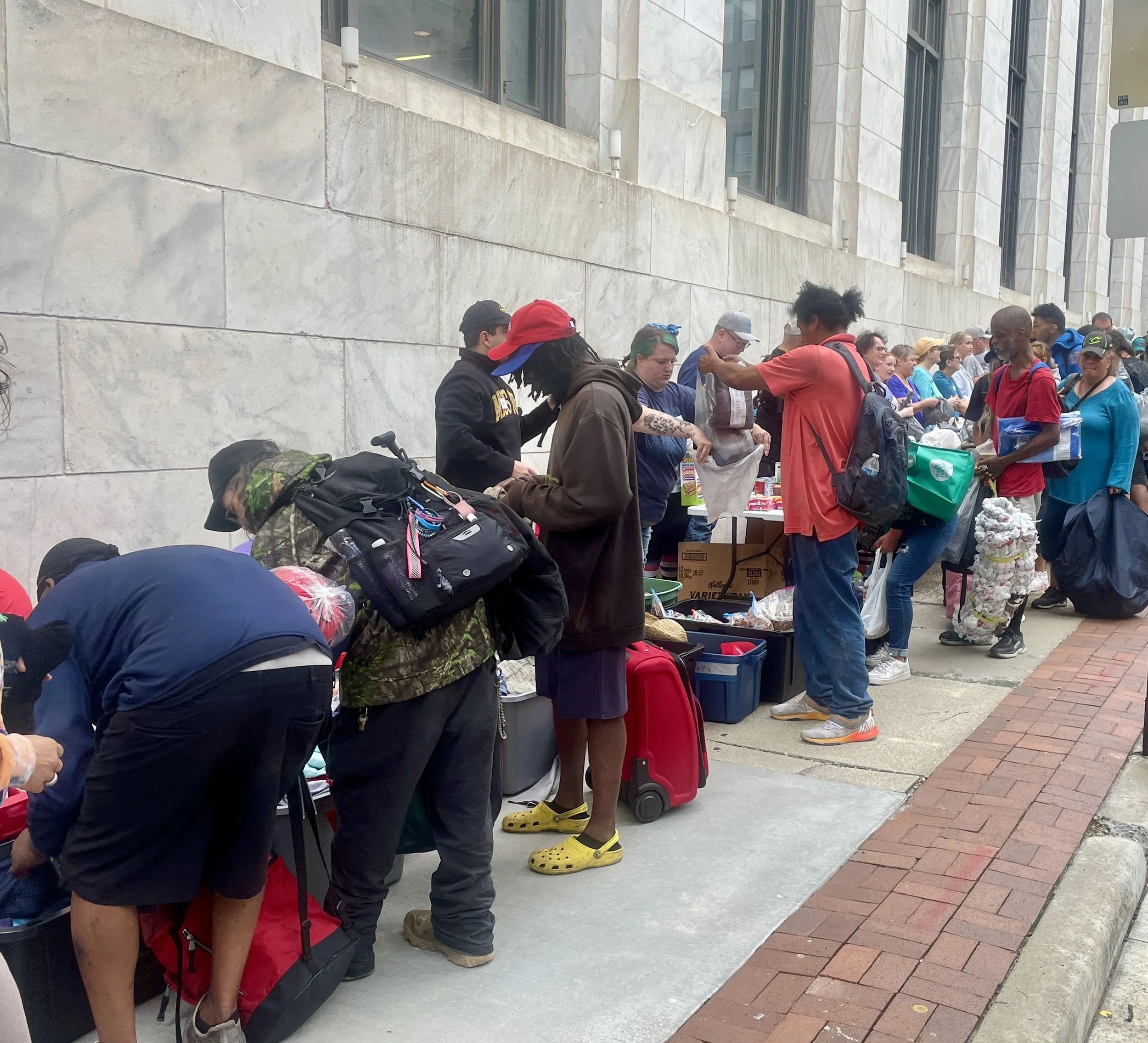 People forming a line outside a building with a table of supplies and backpacks.