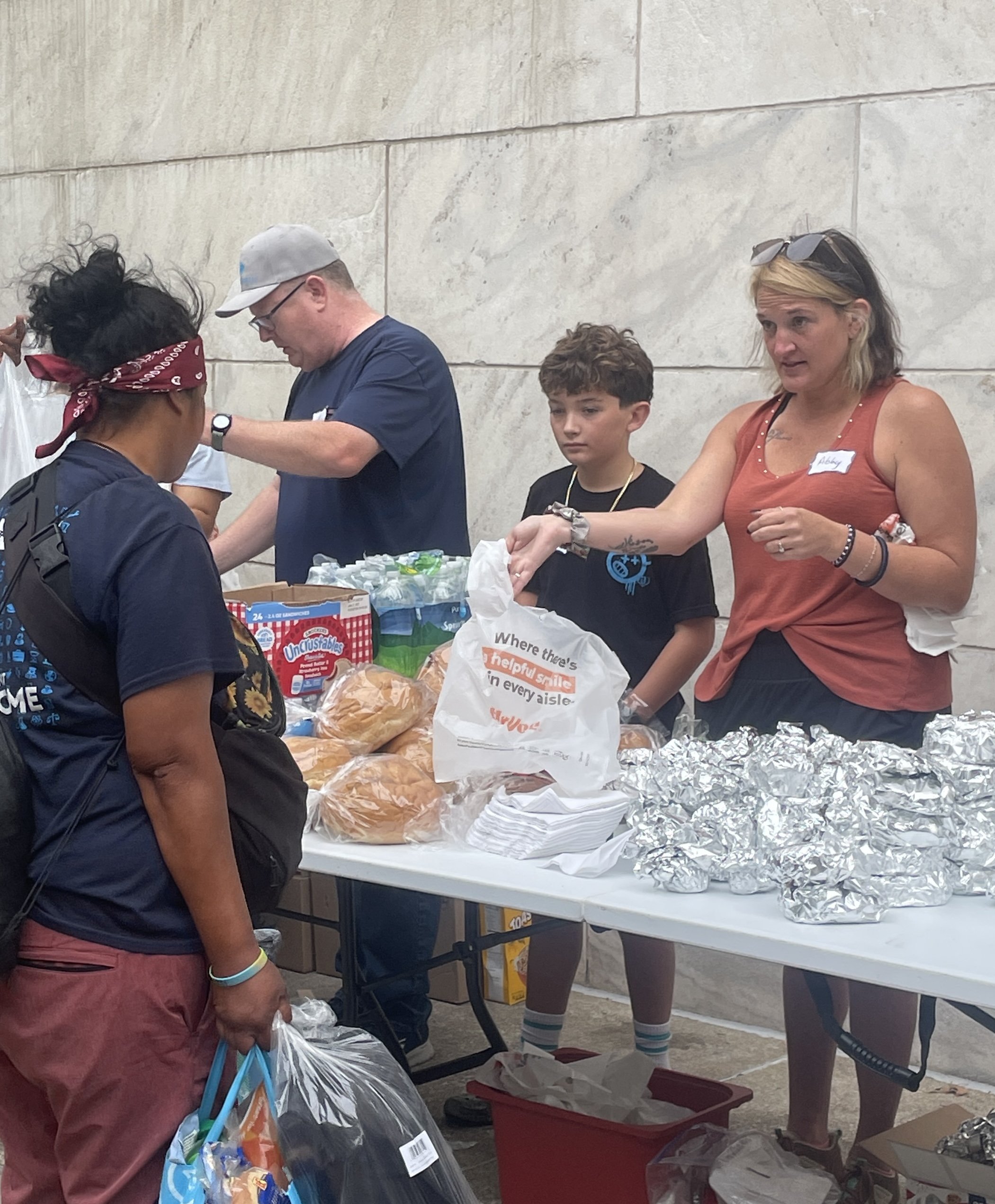 People standing at a table with baked goods, including bread, wrapped in plastic or foil, in an indoor setting with a concrete wall background.