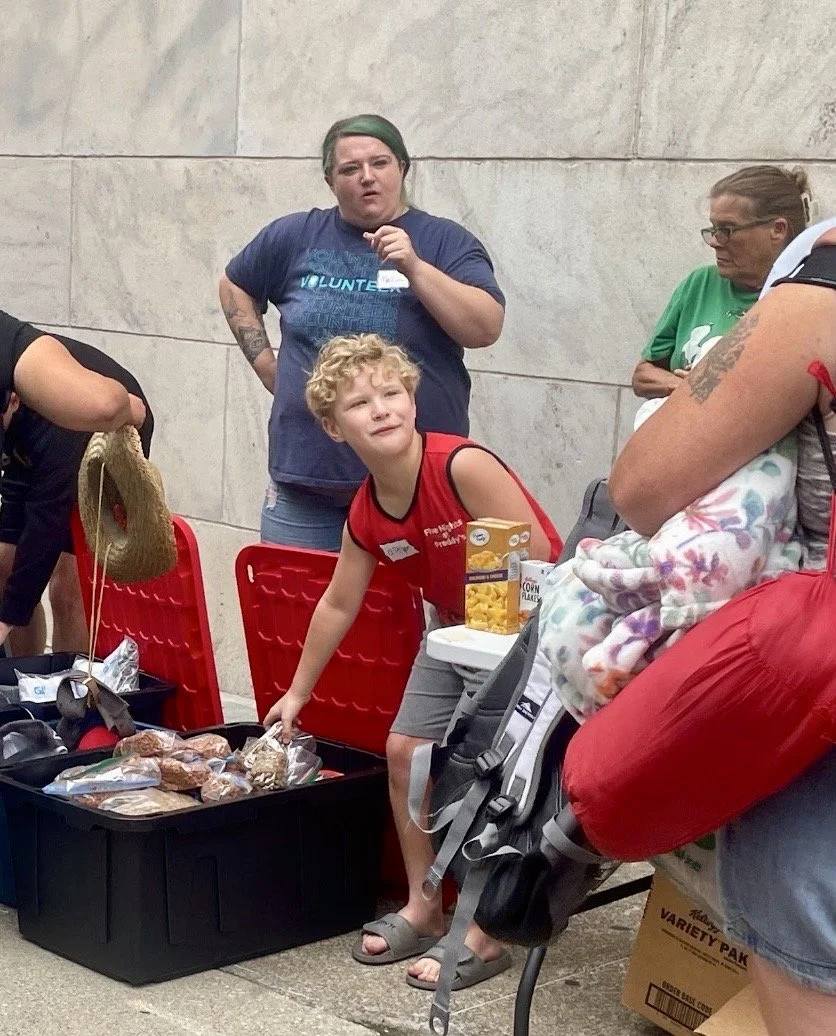 A young boy with curly blonde hair standing next to a red shopping cart filled with snacks, looking up and talking to a person carrying a blanket. Several women are standing behind him, one wearing a volunteer shirt and another with glasses and a gre