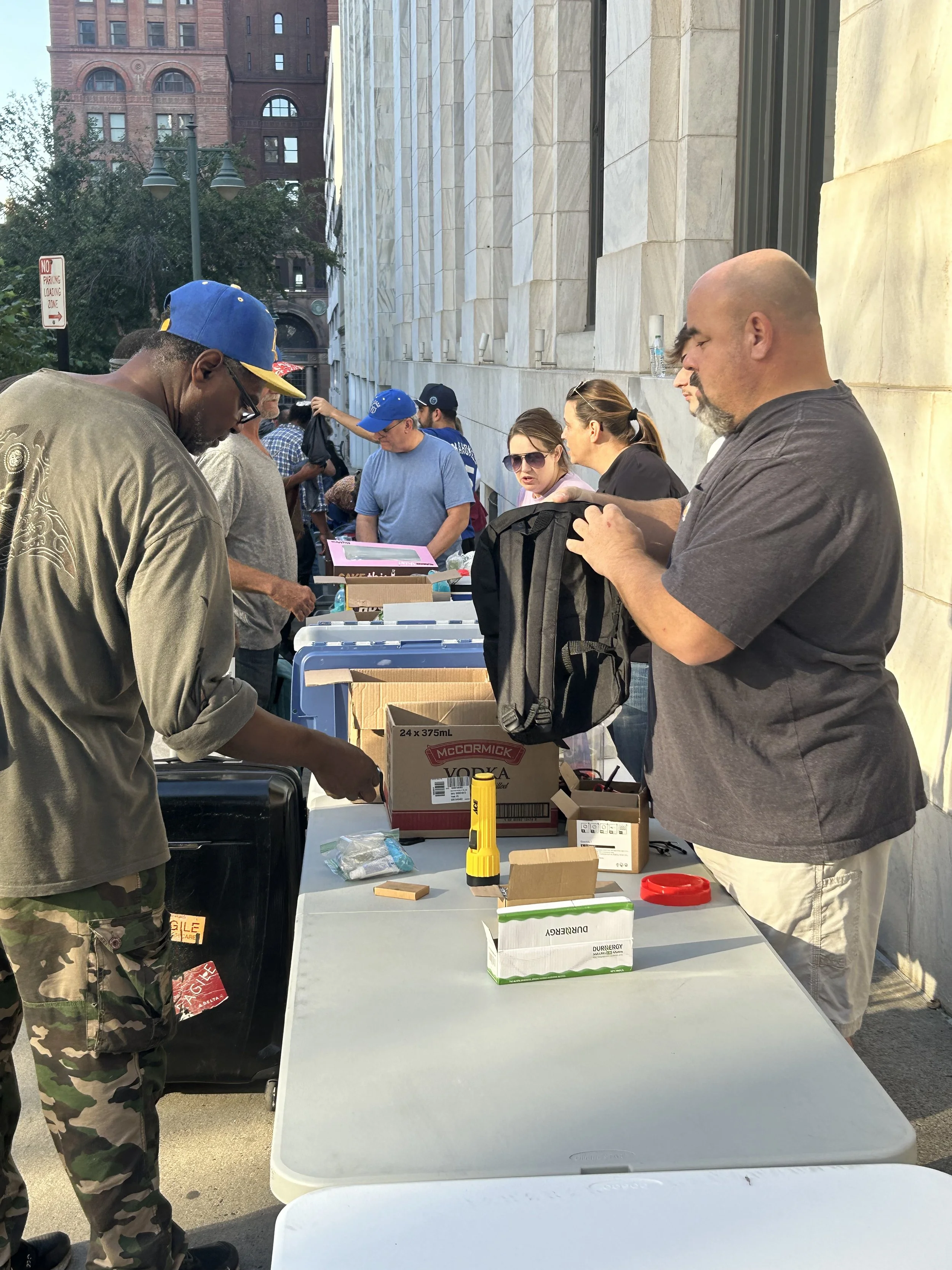 People standing in line at an outdoor event, with tables and boxes on the table, on a city street with buildings in the background.