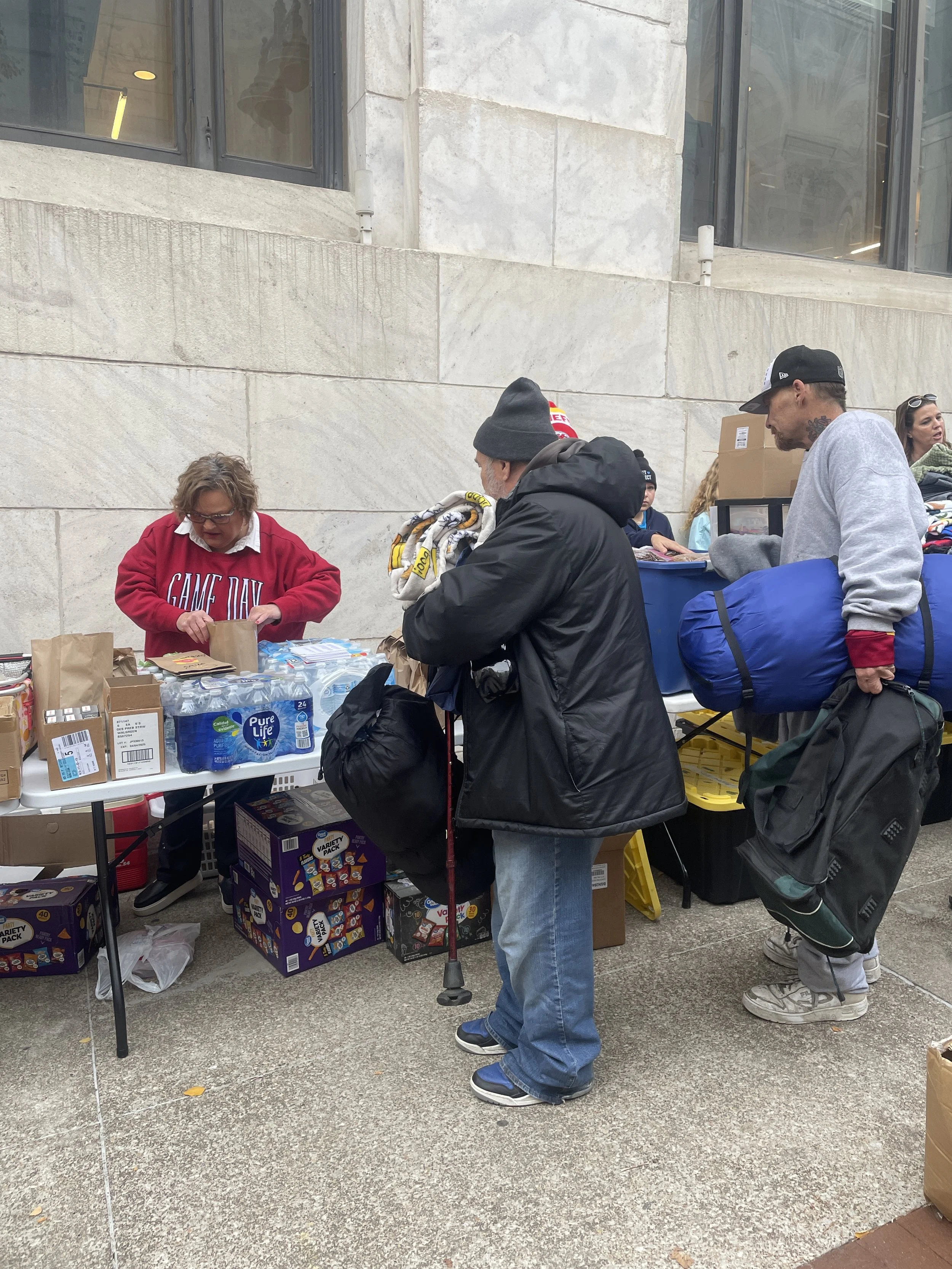 People at a Street Support KC food distribution table with bottled water, snacks, and supplies outside a building.