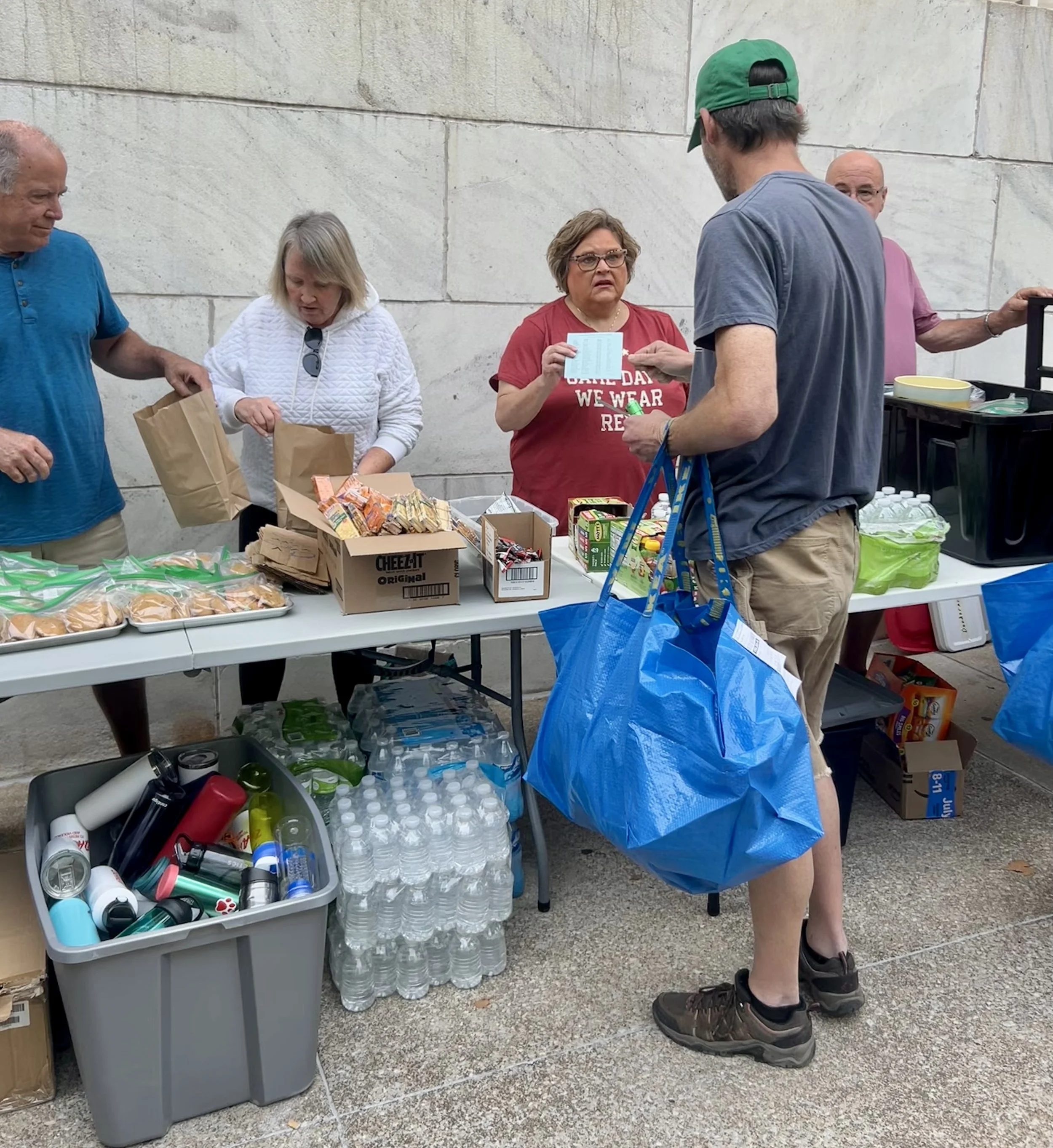 People serving food and drinks at a community event table for Street Support KC, with food packets, bottled water, and supplies.