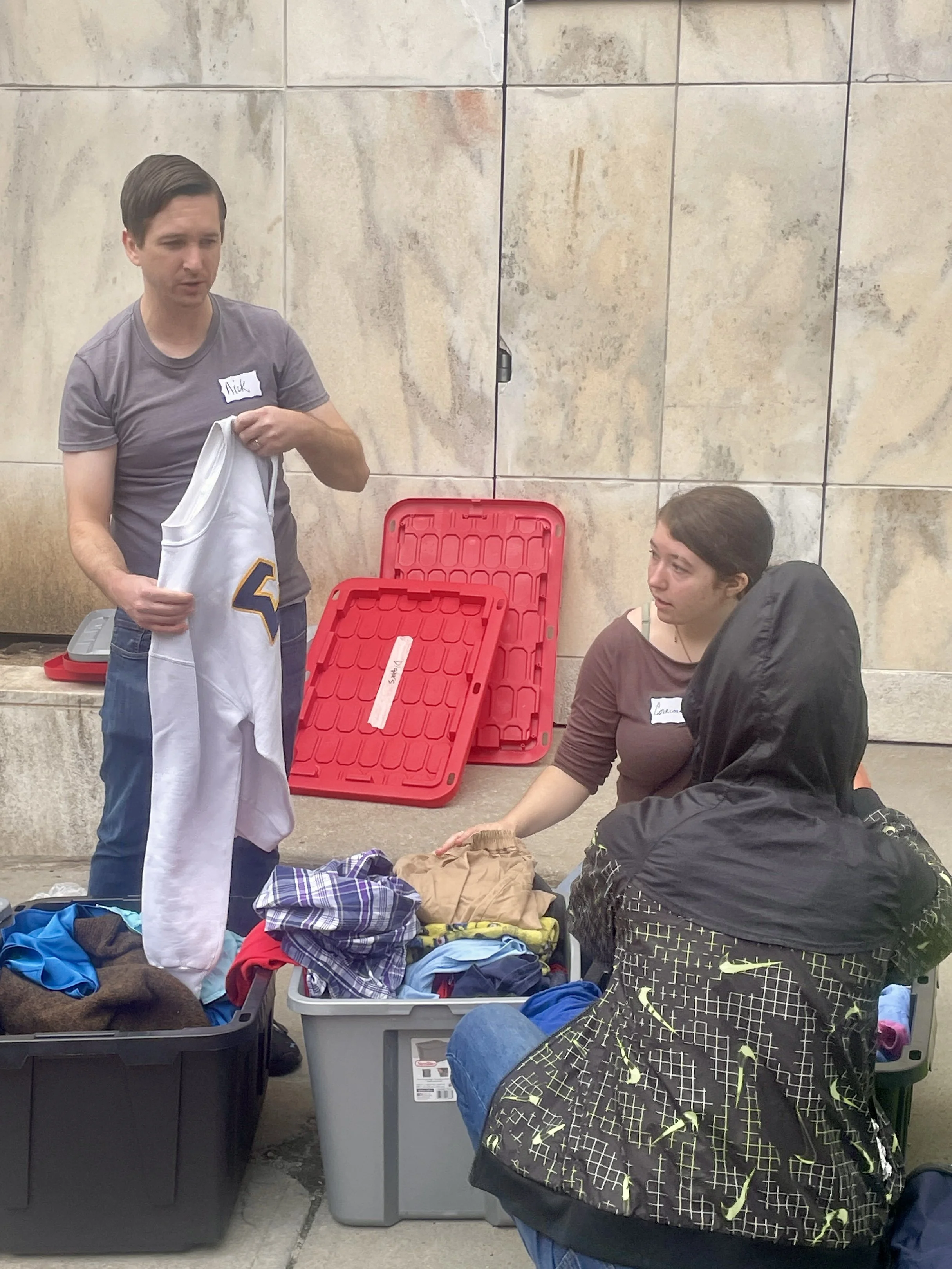 People sorting through clothing and other items in large storage bins, with some wearing name tags, outdoors against a concrete wall.