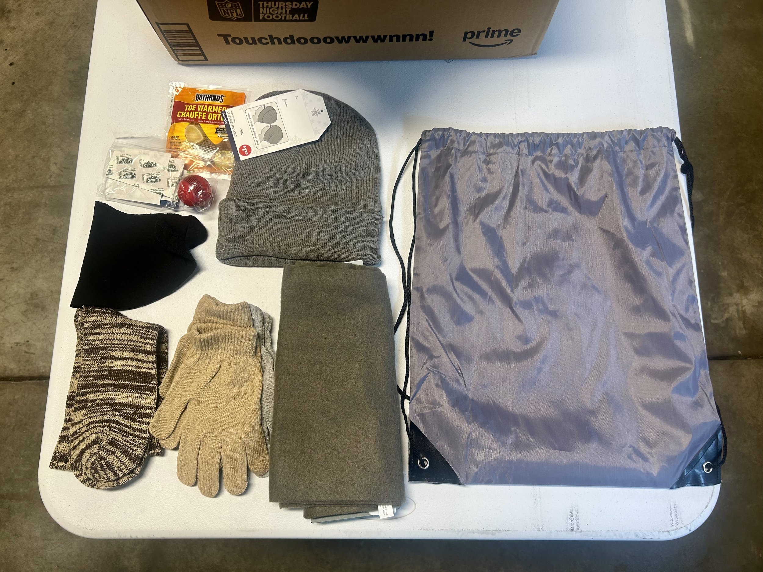 A collection of outdoor winter gear and accessories on a white table, including gloves, hats, socks, and a gray drawstring bag, with a cardboard box in the background.