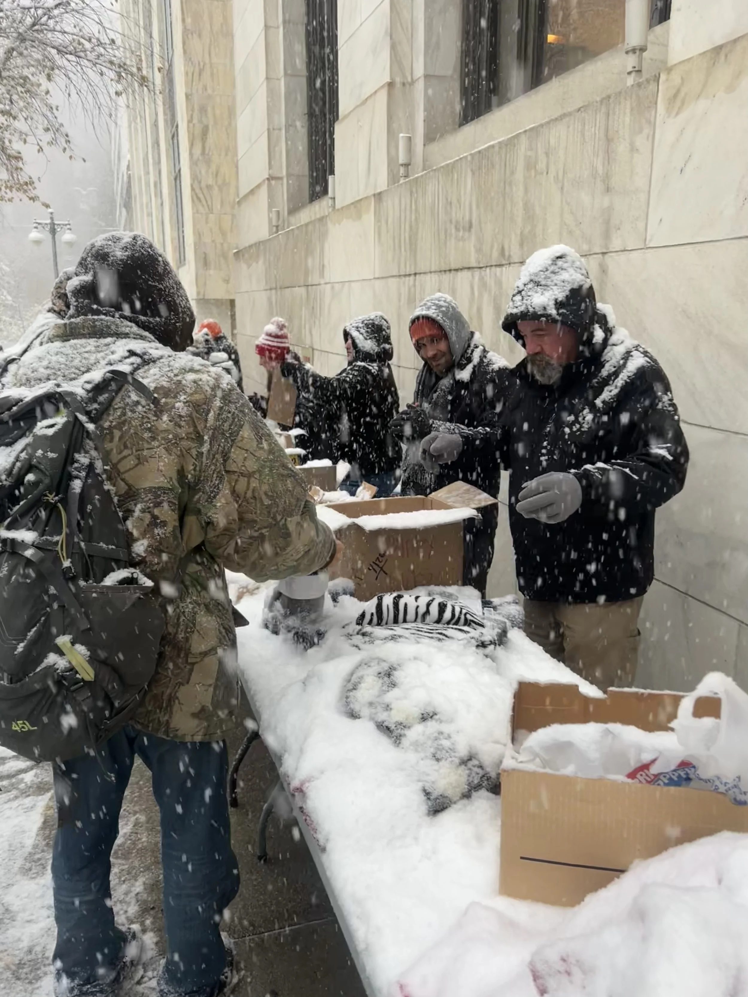 Group of people in winter clothing handing out food and supplies from boxes on a table outside in snowy weather for Street Support KC