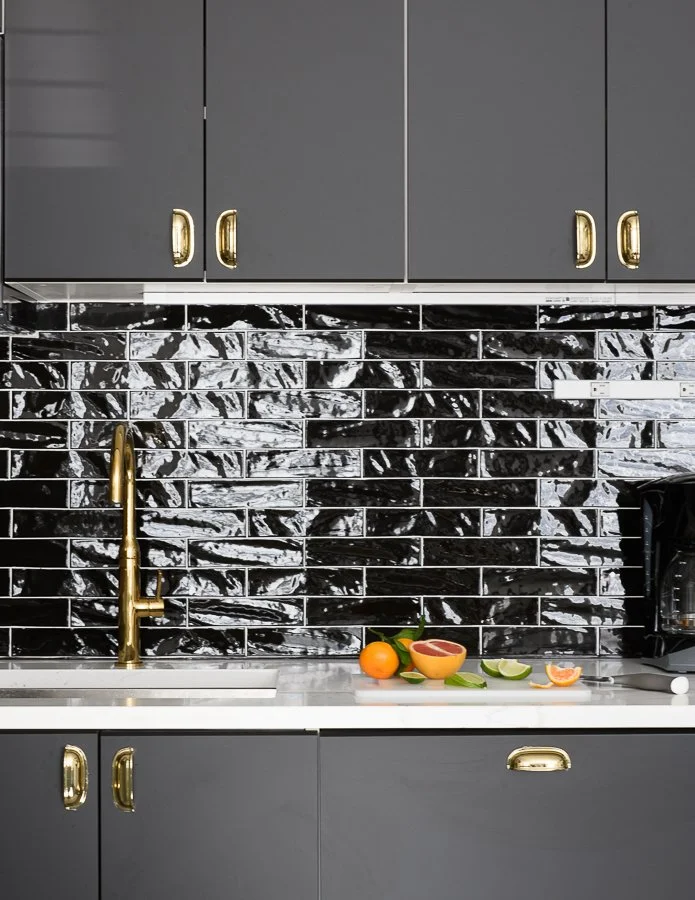 Modern kitchen with grey cabinets, black marble backsplash, gold faucet, and a white countertop with sliced citrus fruits and a grapefruit on it.
