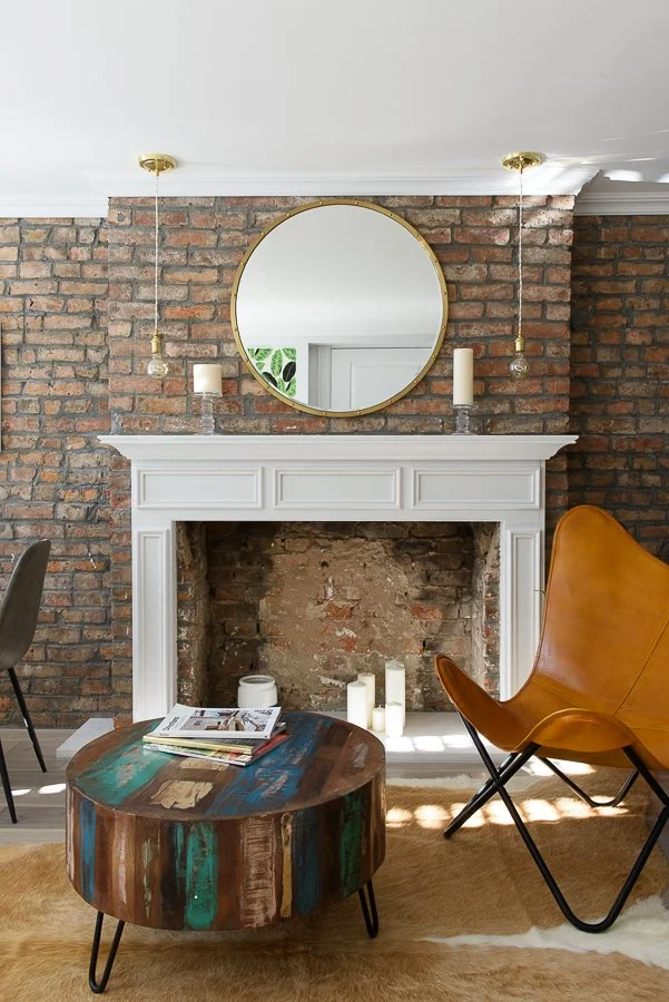 Living room fireplace with a white mantel against a brick wall, decorated with candles and a round mirror, with a leather chair, a colorful wooden coffee table, and reading materials.