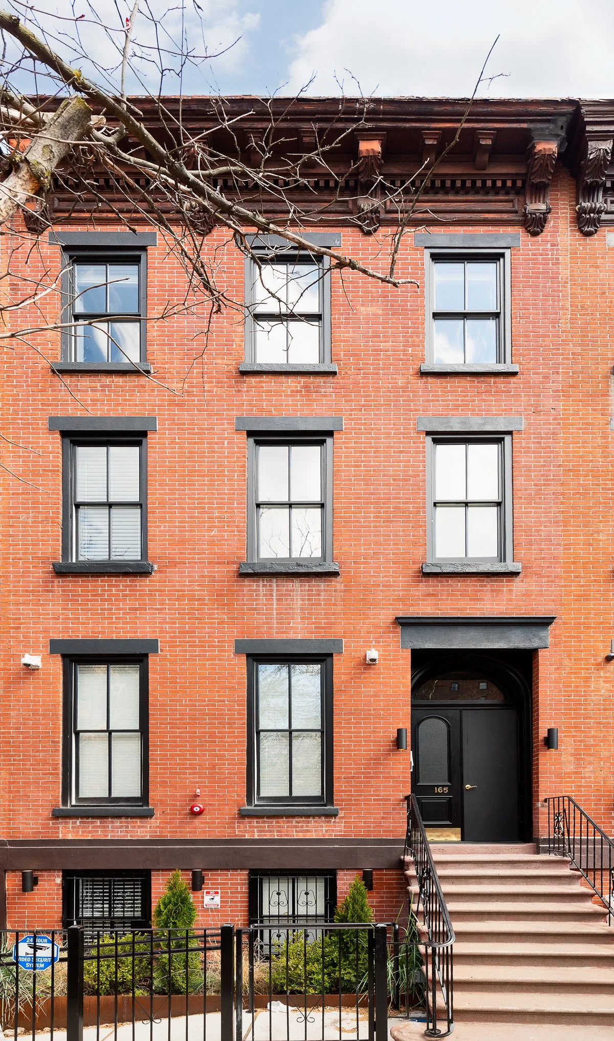 A brick apartment building with black-framed windows, a black door, and stairs leading to the entrance. A small yard with shrubs is in front, and a leafless tree branch extends across the top of the image.