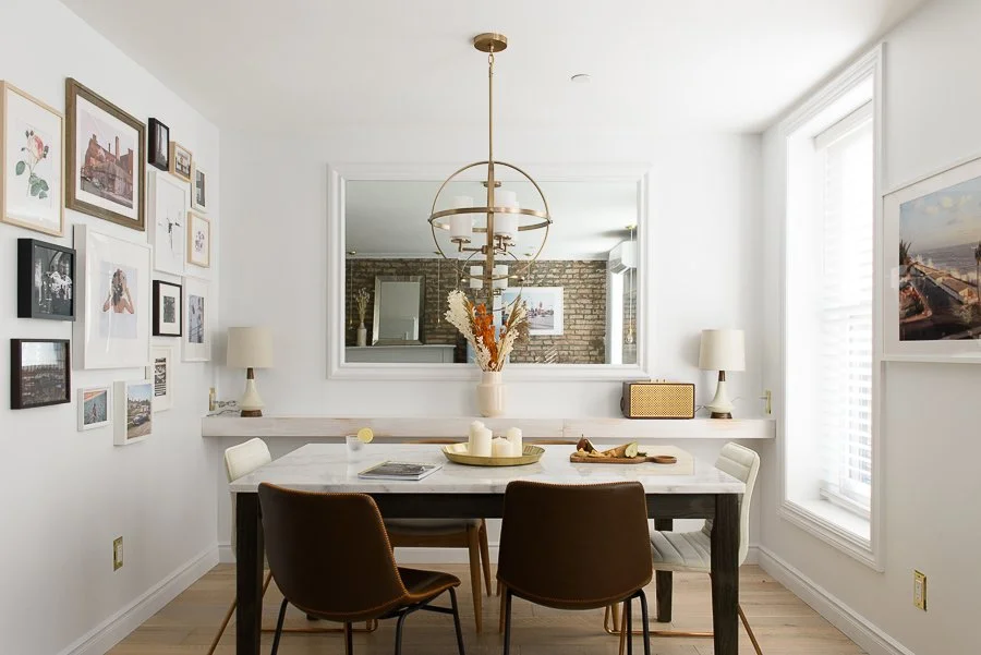 Dining room with six chairs, a white table with candles and decorative items, wall-mounted mirrors, framed art, and windows with blinds.