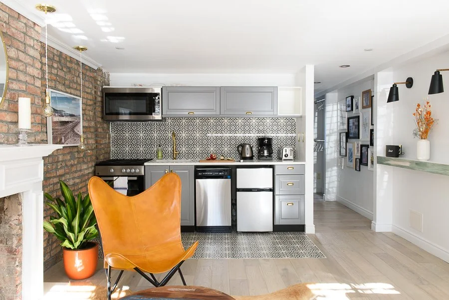 A modern kitchen with gray cabinets, patterned backsplash, stainless steel appliances, and an orange leather chair near a green potted plant, brick wall, and gallery wall of framed photos.