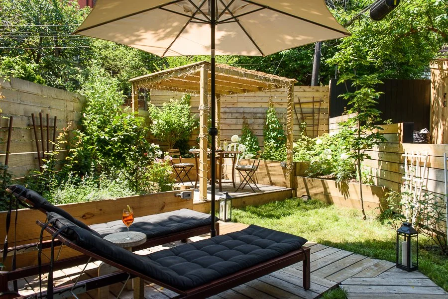 Backyard patio with a wooden lounge chair, small side table with a drink, large umbrella, lush garden, wooden deck, and a small covered seating area with outdoor table and chairs.