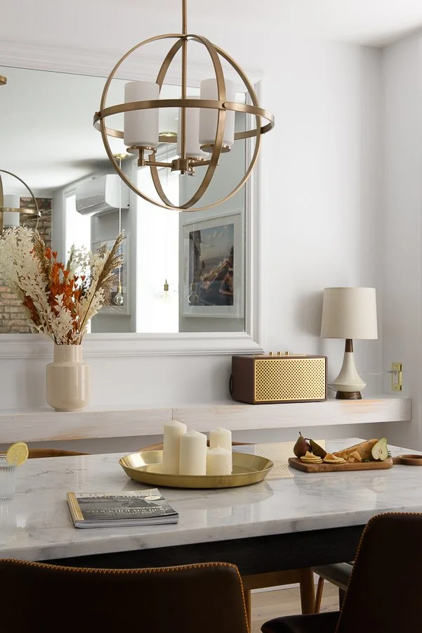 A modern dining room with a white marble table, brass candle holder with white candles, a vase with dried flowers, a table lamp, a decorative speaker, and a mirror on the wall reflecting part of the room.