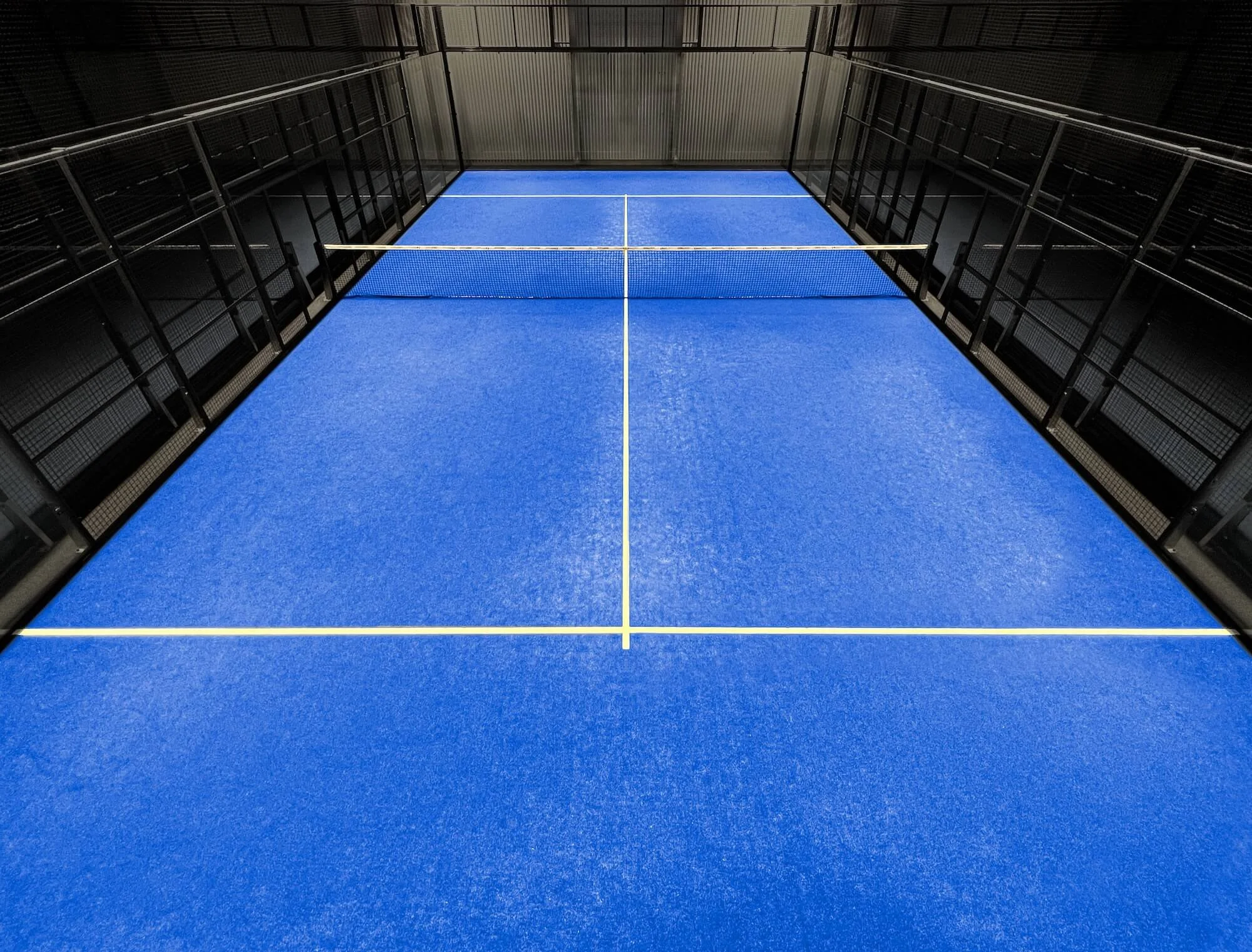 Leerer Padelplatz mit blauer Oberfläche und weissen Linien, umgeben von schwarzen Gitterzäunen, im Inneren einer Halle.