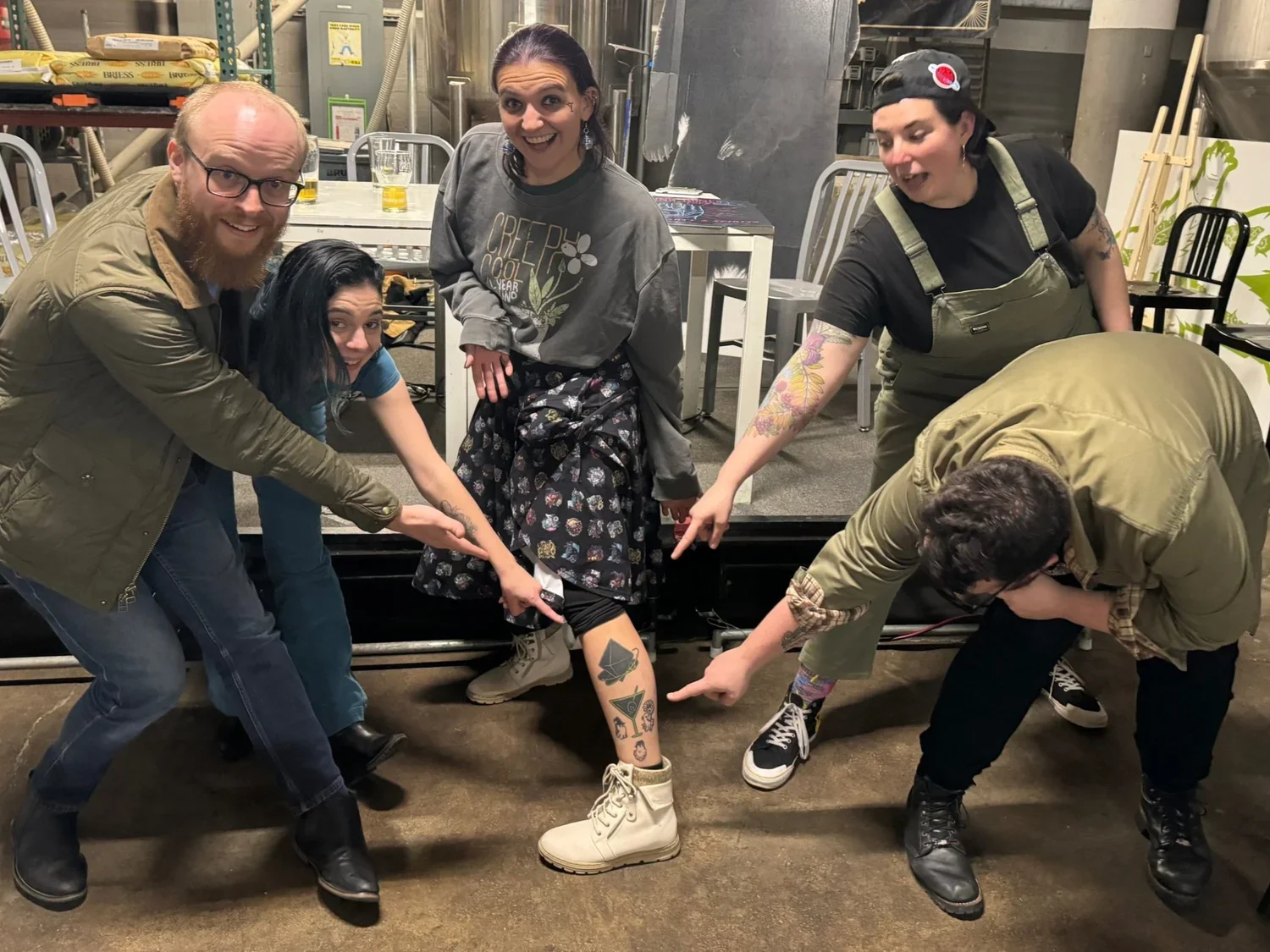 Five people in a restaurant or bar, pointing at a woman's tattooed leg, which has multiple tattoos including a rocket, a cat, and a face. One woman is holding a knife, and everyone is smiling or laughing.
