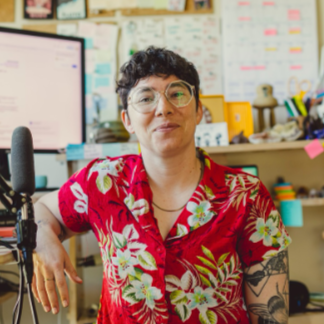 A person with dark curly hair, glasses, and a bright red patterned shirt sits in a warm and colorful office with a microphone behind them