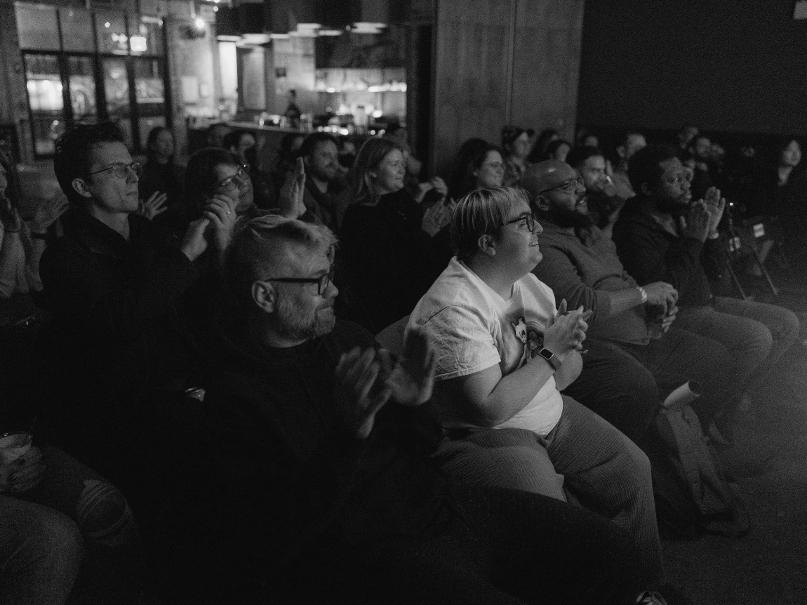 Audience members sitting in a darkened space, watching a performance or presentation and clapping.