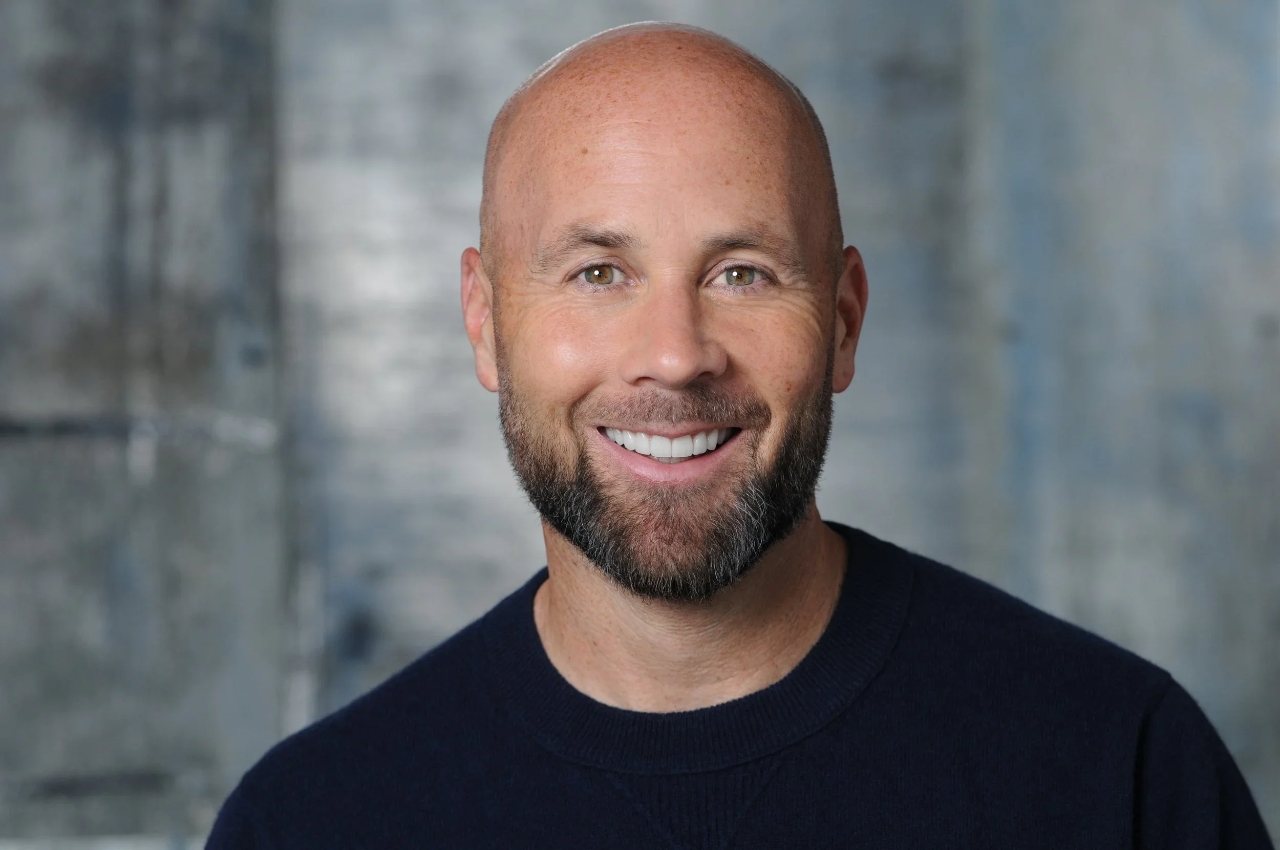 A photo of a white man with a short beard smiling against a gray/blue background