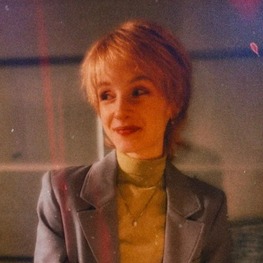 Close-up of a person with short blonde hair, a green turtleneck, and a gray blazer sitting against a wall with light gray paneling
