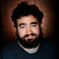 A smiling man with black curly hair and a full beard, against a brown background wearing a gray shirt and black sweater