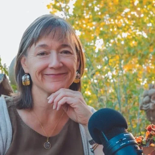 A woman with shoulder-length brown and gray hair smiling in front of trees and flowers, with a microphone and gardening shears in front of her