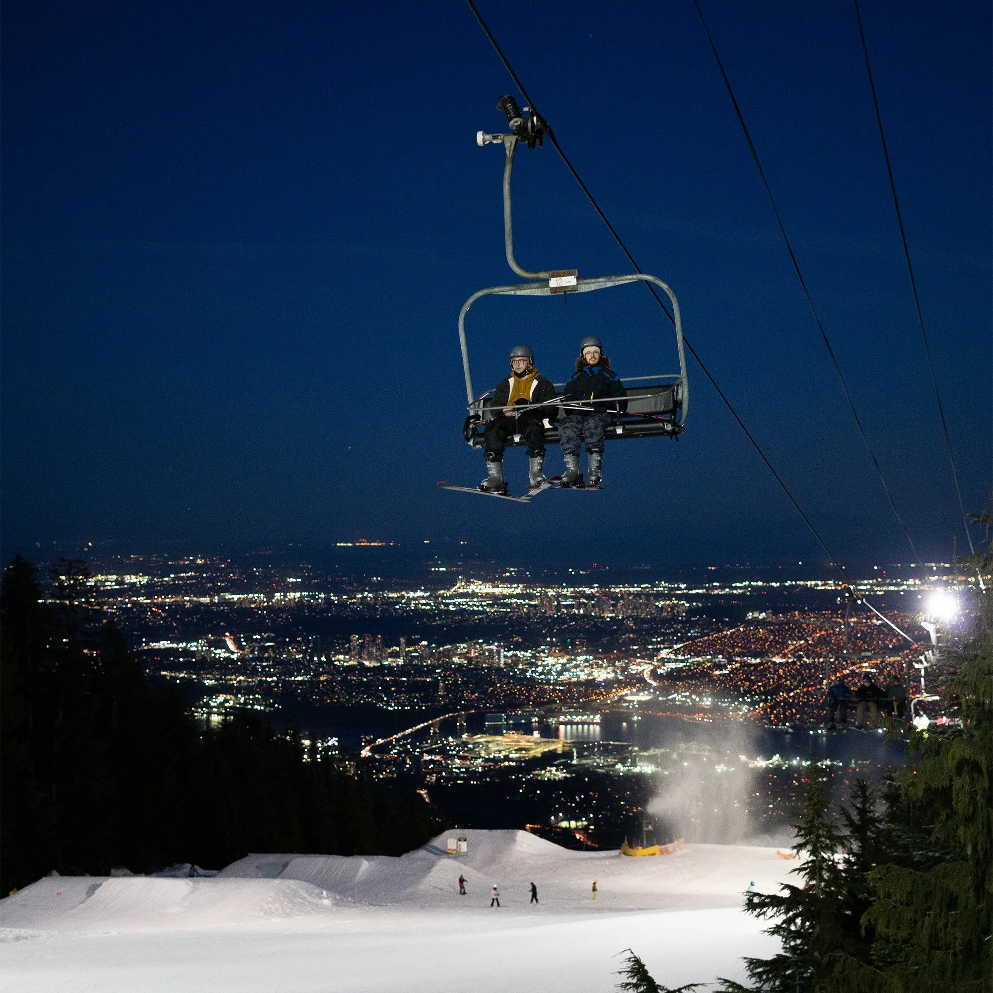 Two skiers sitting on a ski lift at night above a cityscape with snow, trees, and illuminated city lights.