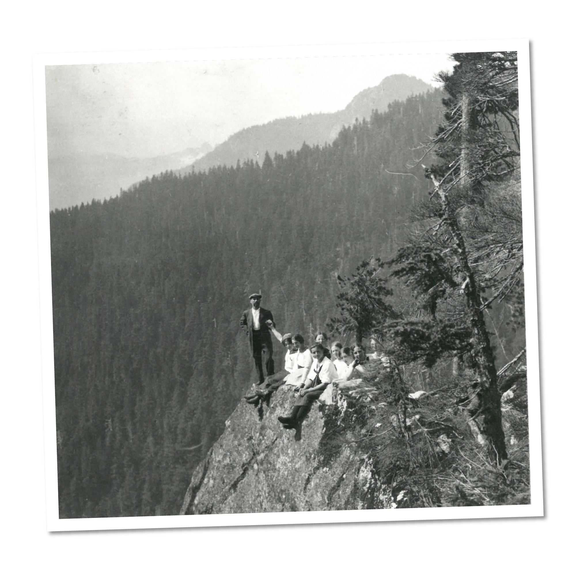 Black and white photo of a man standing and five children sitting on a rocky cliff with a mountainous forest background.
