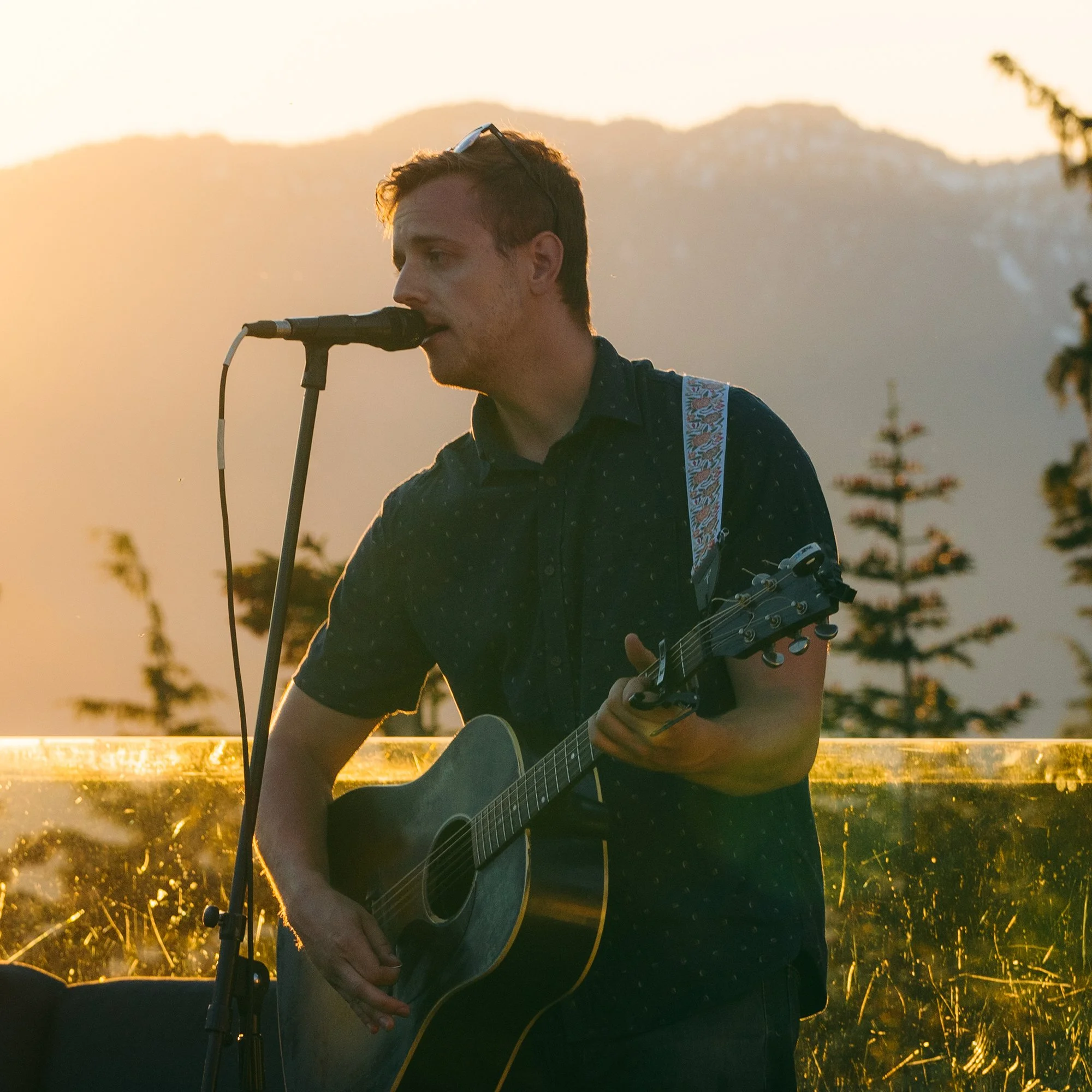 A man with glasses on his head playing an acoustic guitar and singing into a microphone during sunset with trees and mountains in the background.