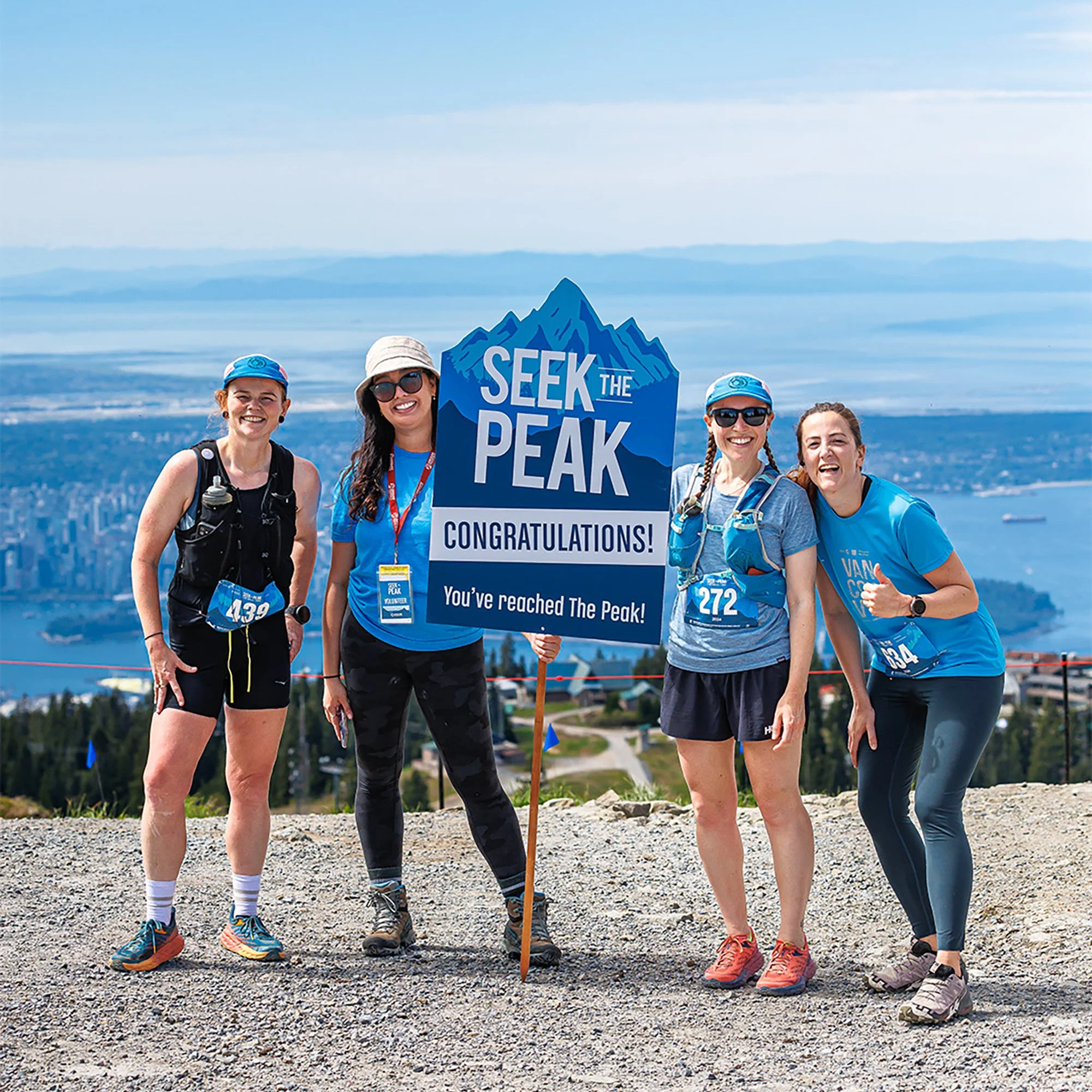 Four women celebrating at the summit of a mountain, holding a sign that reads 'Seek the Peak, Congratulations! You've reached The Peak', with a city skyline and water in the background during daytime.