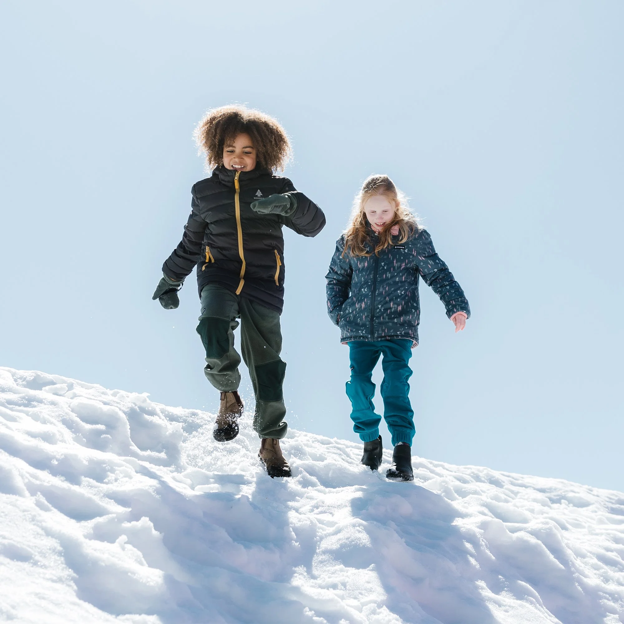 Two children, a girl and a boy, playing in the snow on a sunny winter day, walking down a snowy hill.