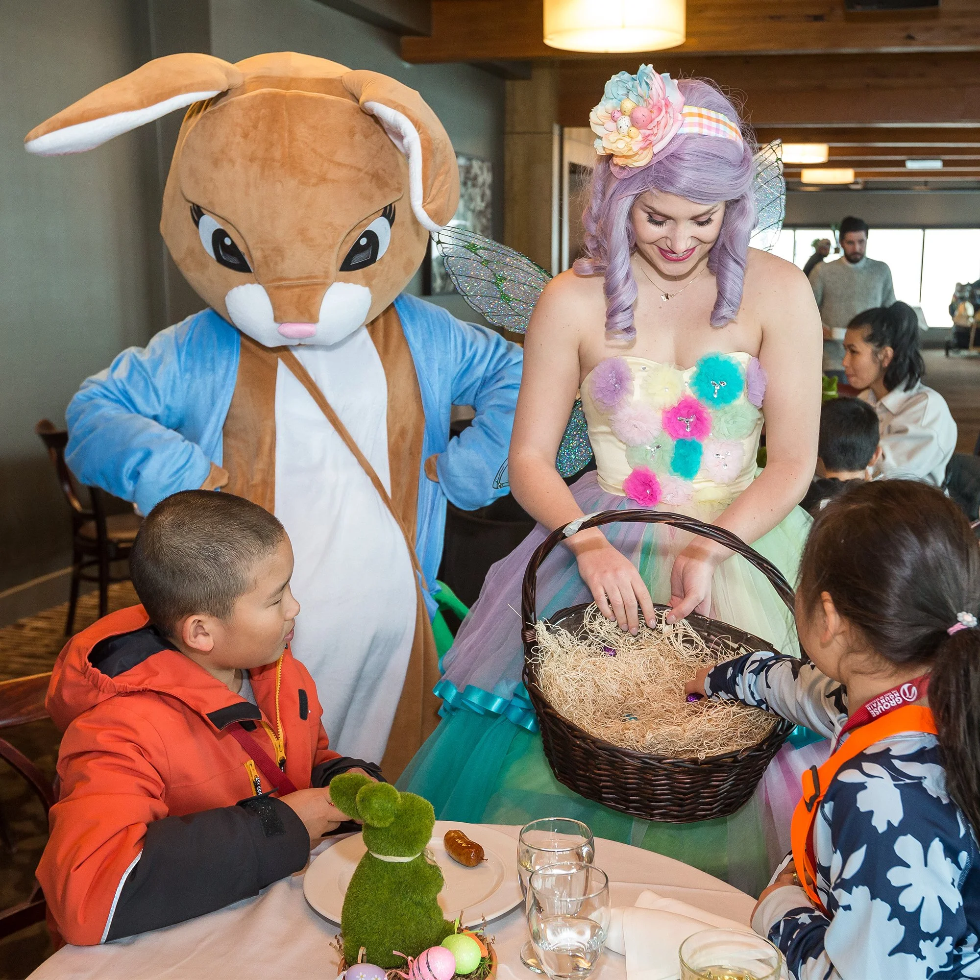 A woman dressed as a fairy in a pastel-colored dress with fairy wings, a person in a bunny costume with large cartoon eyes, and children at a table with Easter decorations and treats, celebrating at an indoor event.