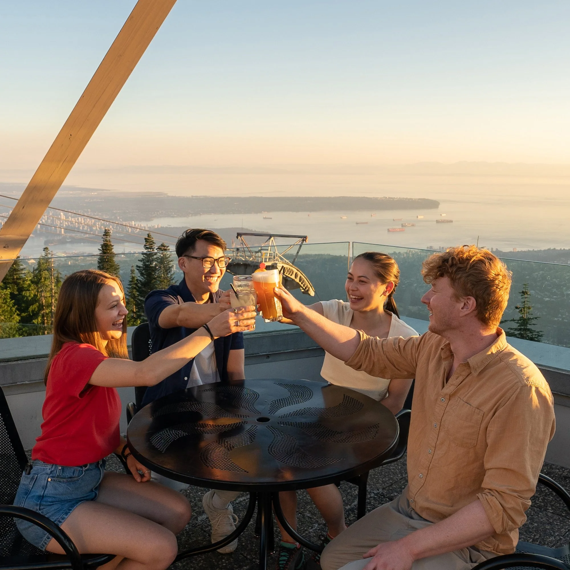 Four friends enjoying drinks and toasting at an outdoor rooftop with city and water view during sunset.
