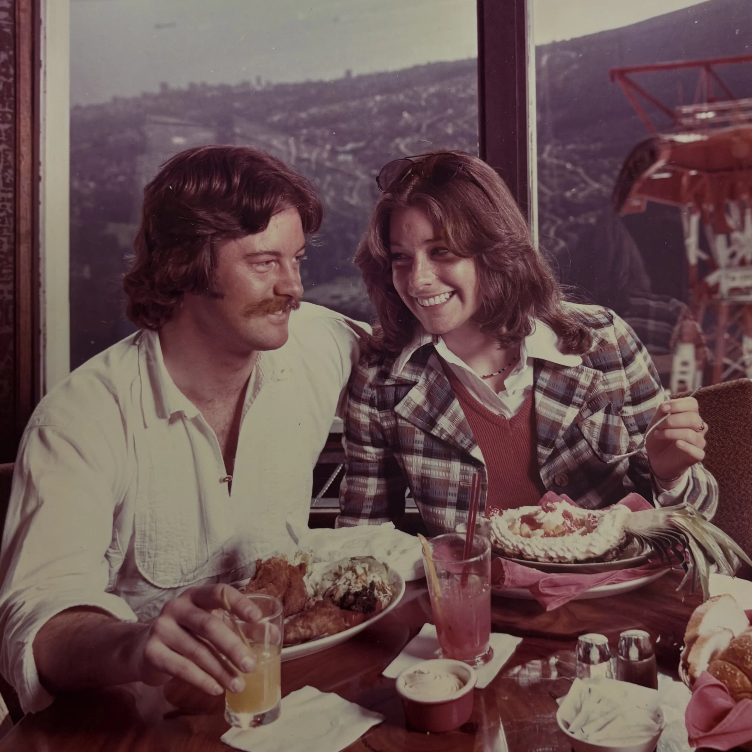 A man and a woman sitting at a table in a restaurant, smiling and sharing a meal, with a city view outside the window.