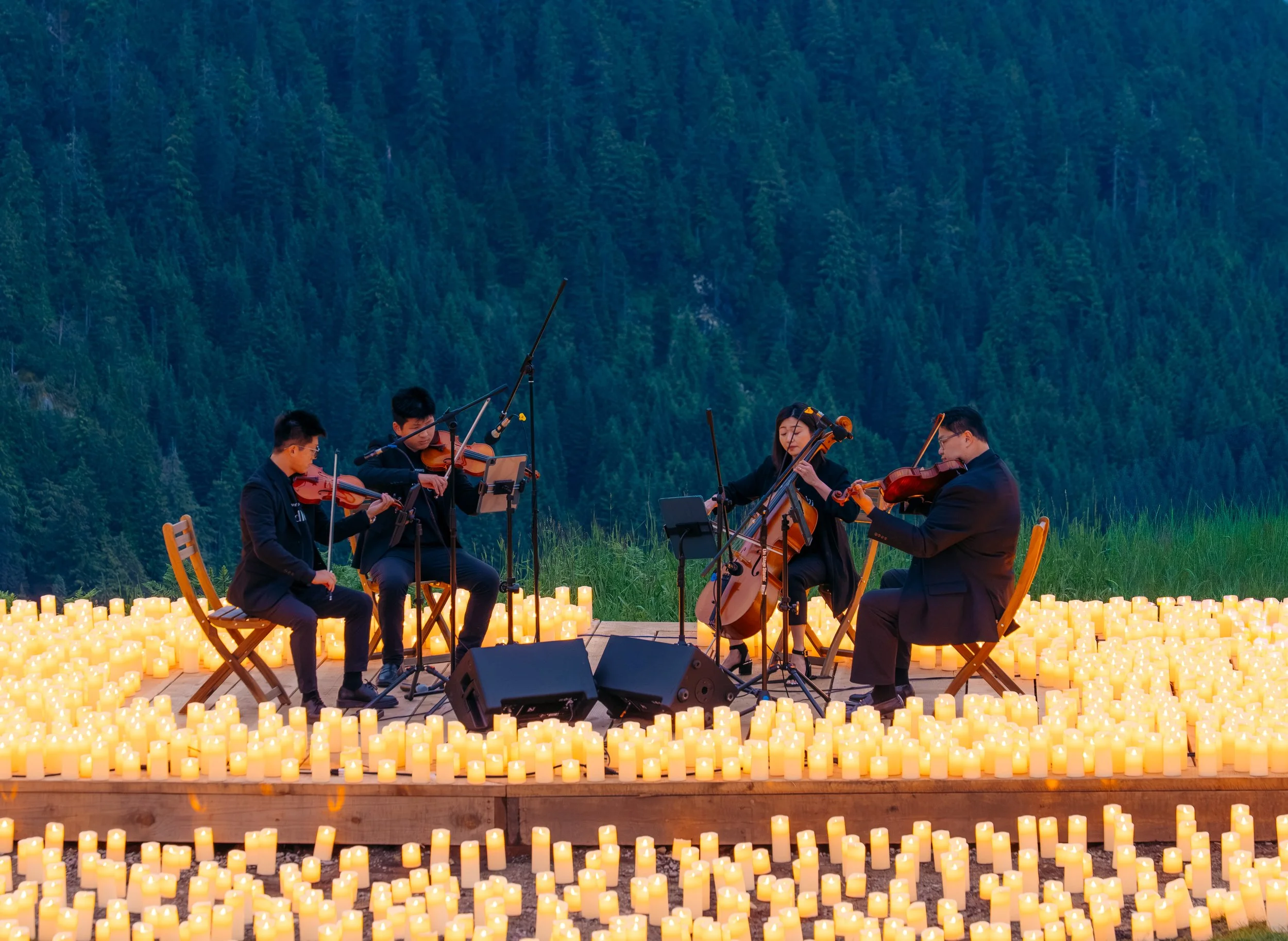 A man with glasses on his head playing an acoustic guitar and singing into a microphone during sunset with trees and mountains in the background.