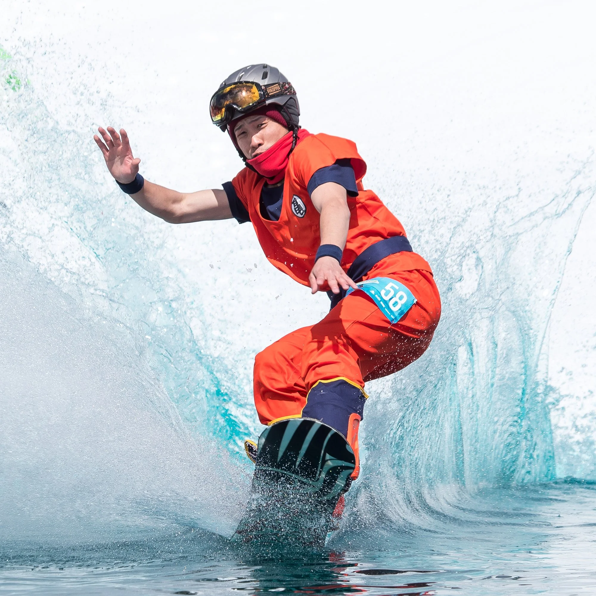 A person snowboarding on a water surface, wearing an orange outfit, a helmet, and goggles, with water splashing around.