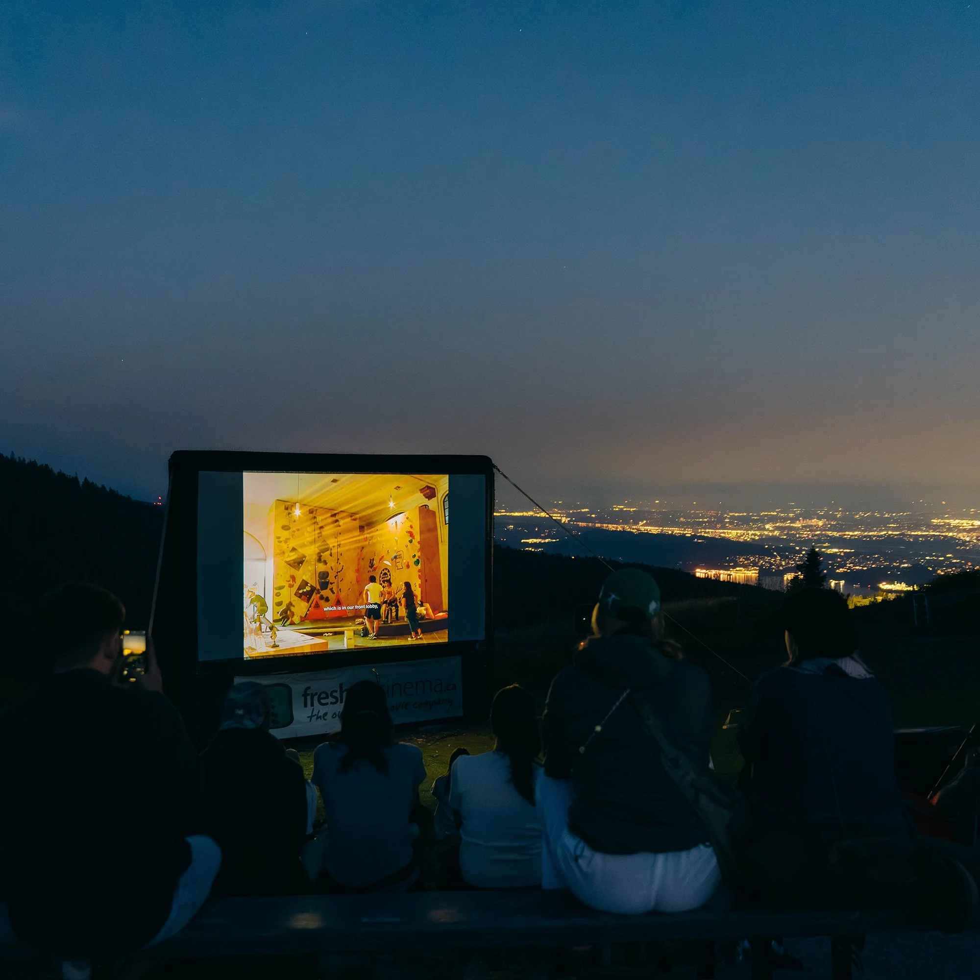 People sitting outdoors watching a movie on a large screen at night, with city lights in the distance.