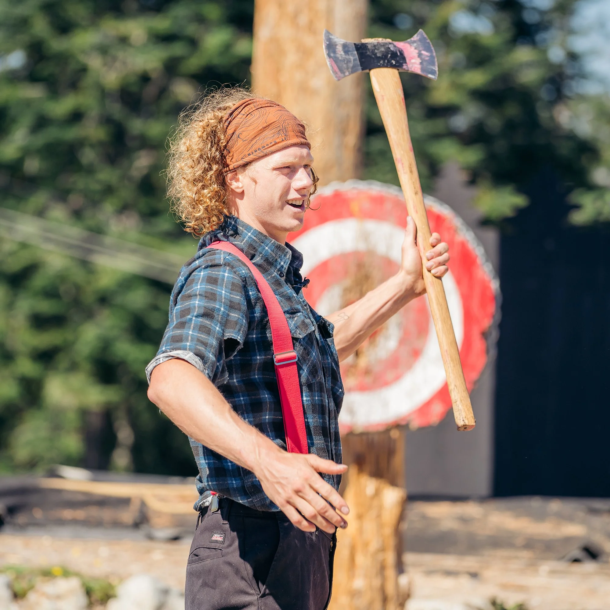 A person with curly red hair holding a large axe, standing outdoors near a wooden target with a red and white circle.