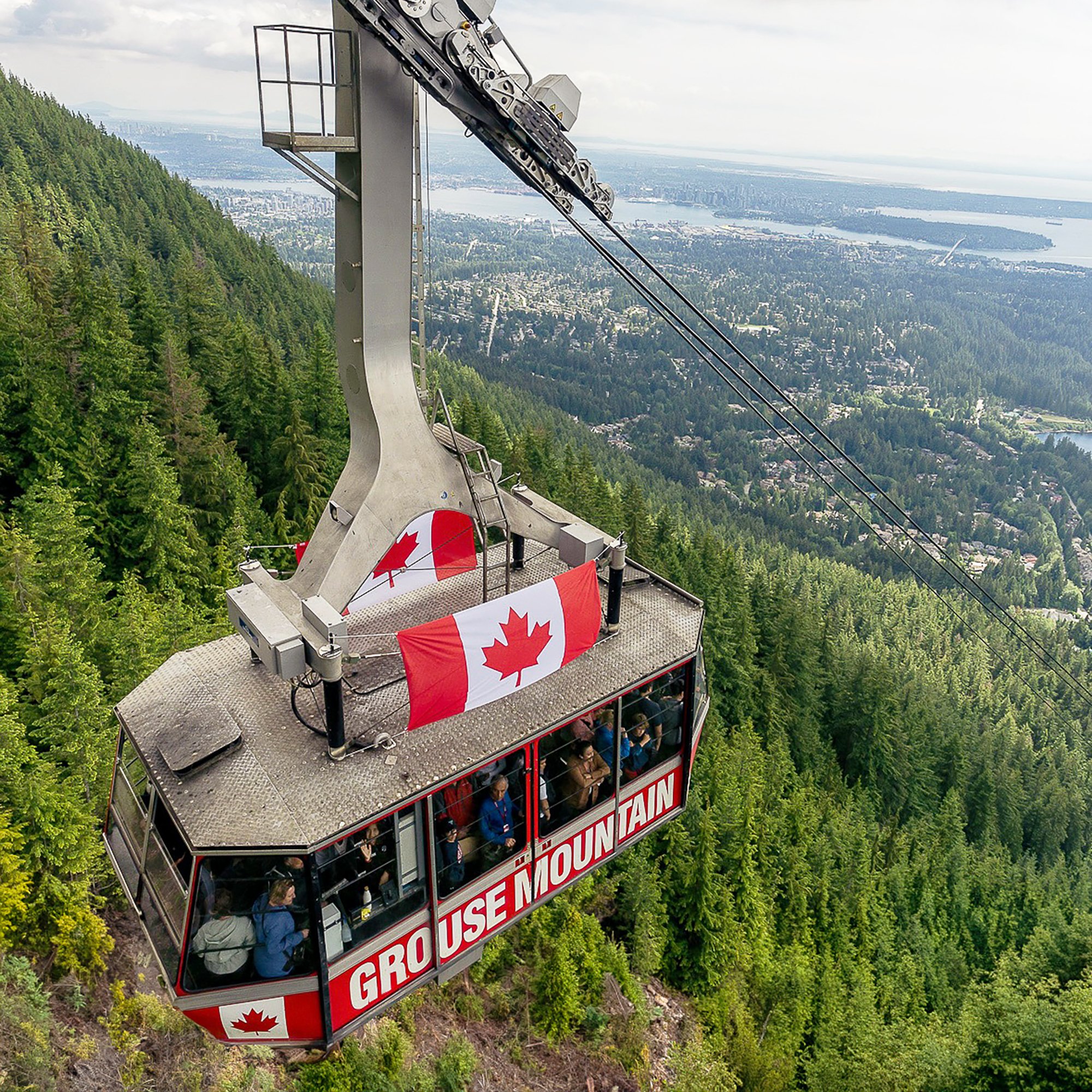 A gondola lift with passengers riding above a forested mountain with a view of a city, water, and distant landscape in the background. The gondola is decorated with Canadian flags and bears the text 'GROUSE MOUNTAIN'.