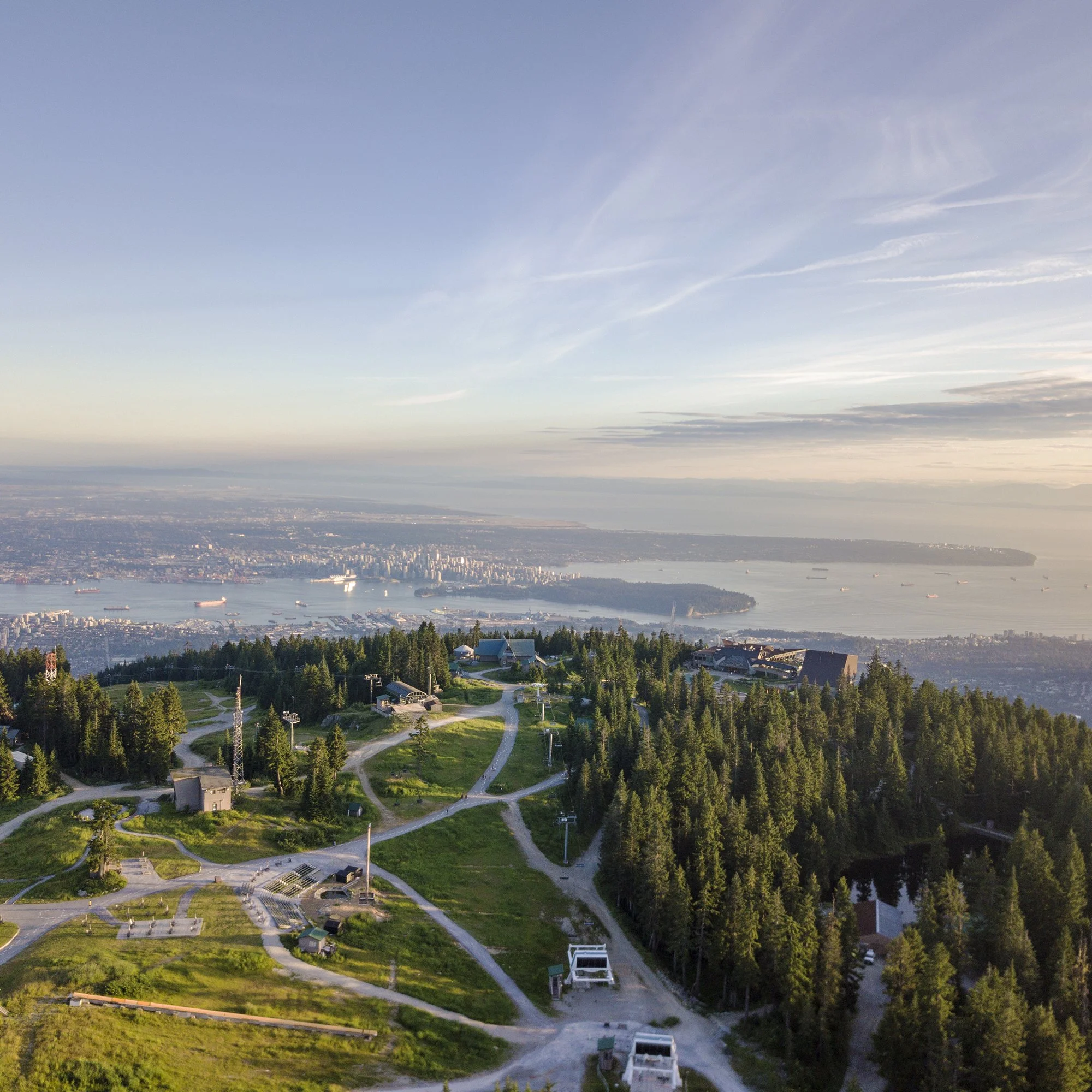 Aerial view of a mountain with a green forest and a few buildings, overlooking a city and harbor in the distance under a partly cloudy sky at sunset.