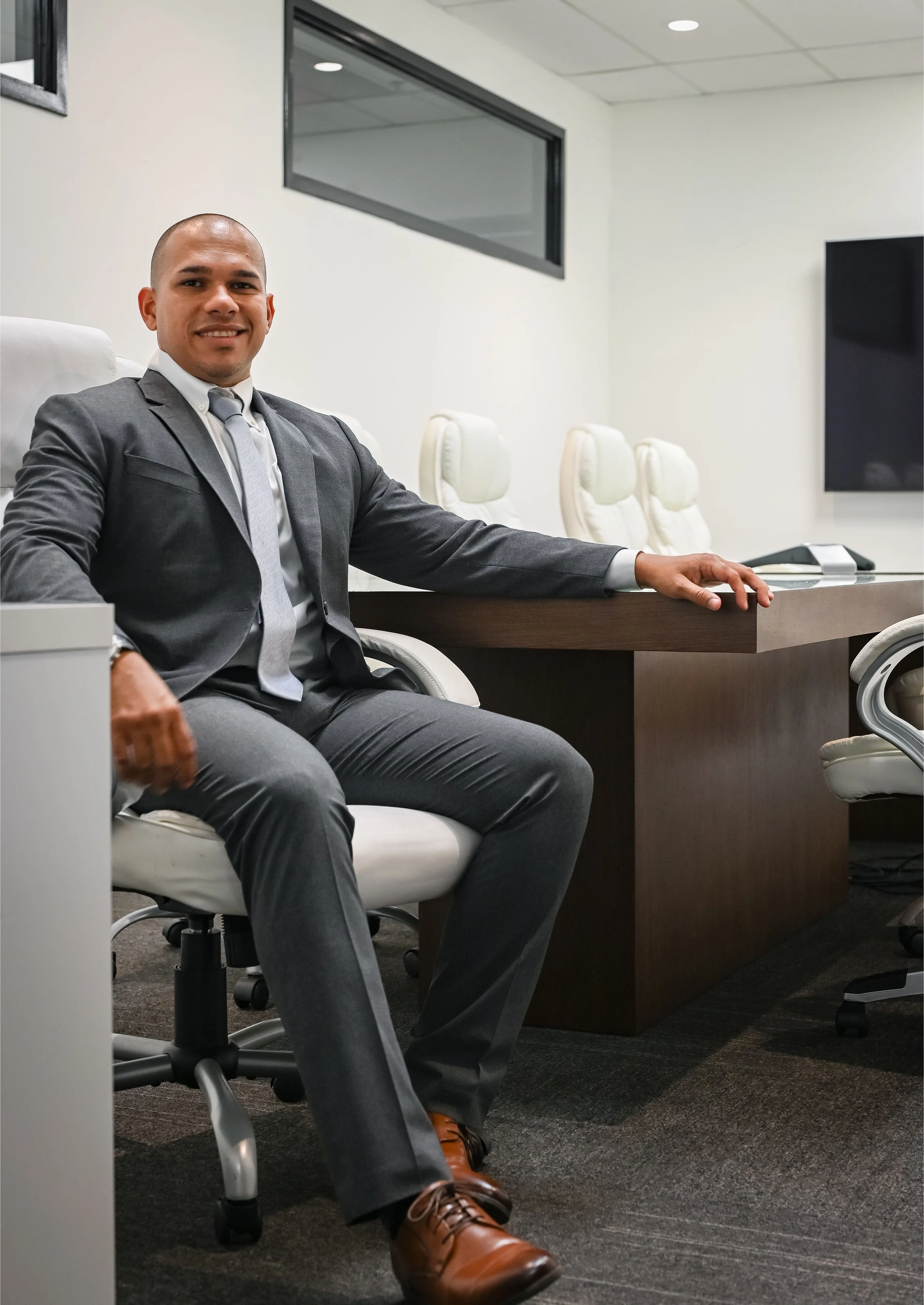 Hombre de negocios sentado en una sala de reuniones con camisa, corbata, traje gris, zapatos marrones, y fondo de sillas blancas y pantallas planas.