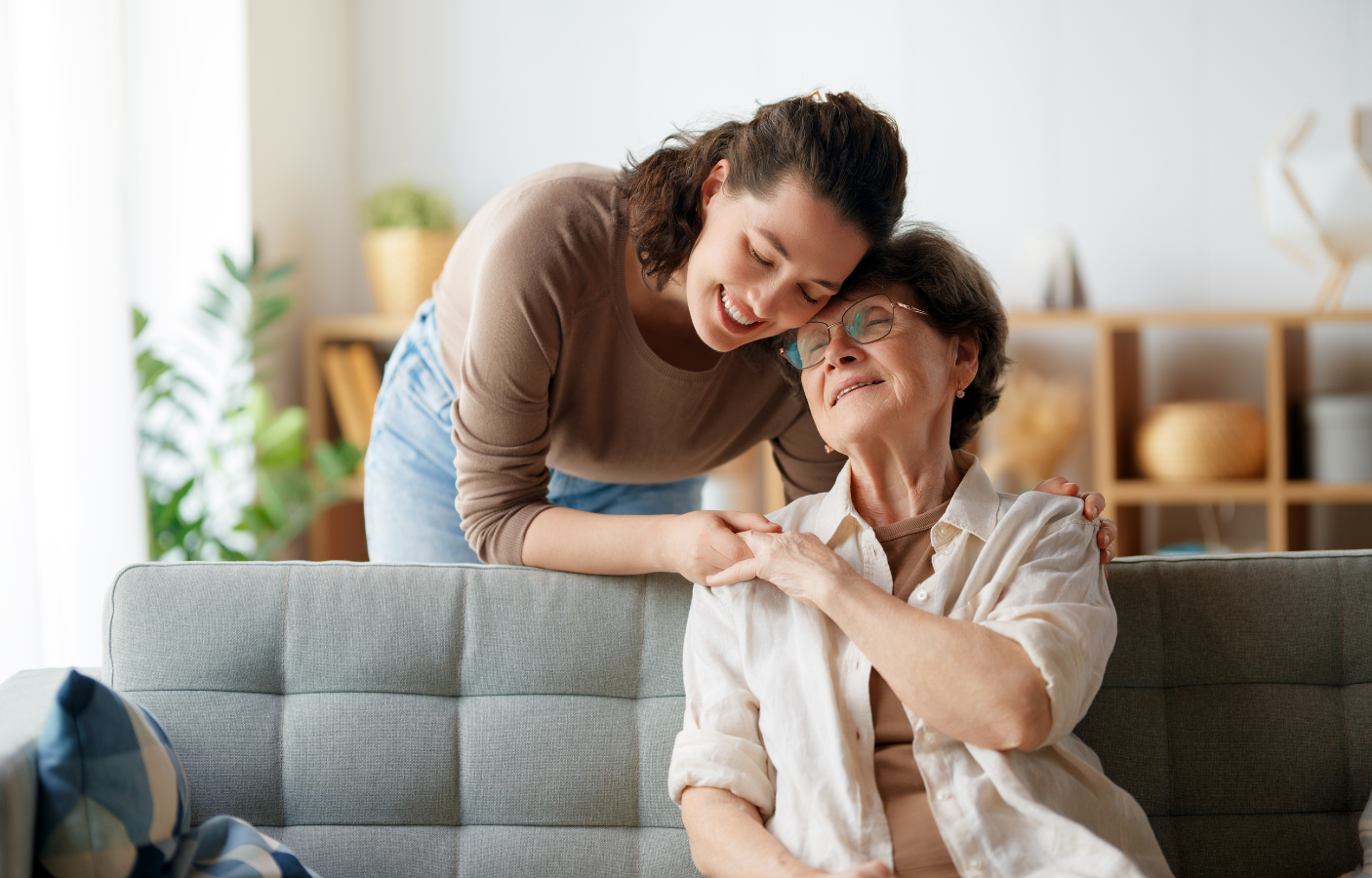 A young woman and an elderly woman sharing a joyful moment on a gray couch in a bright living room.