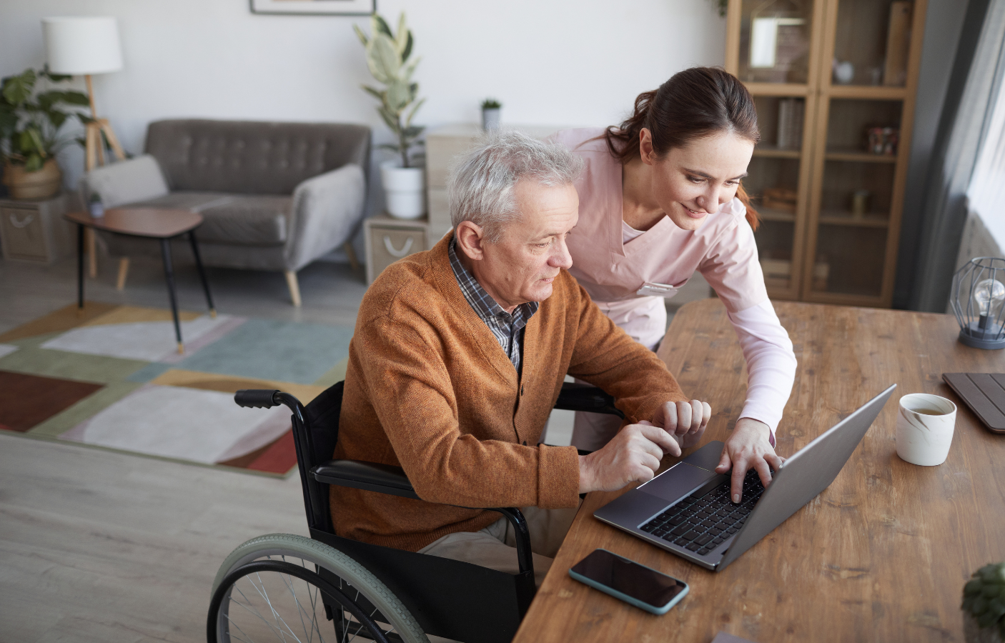 A young female caregiver assisting an elderly man in a wheelchair with a laptop at a wooden table in a living room.