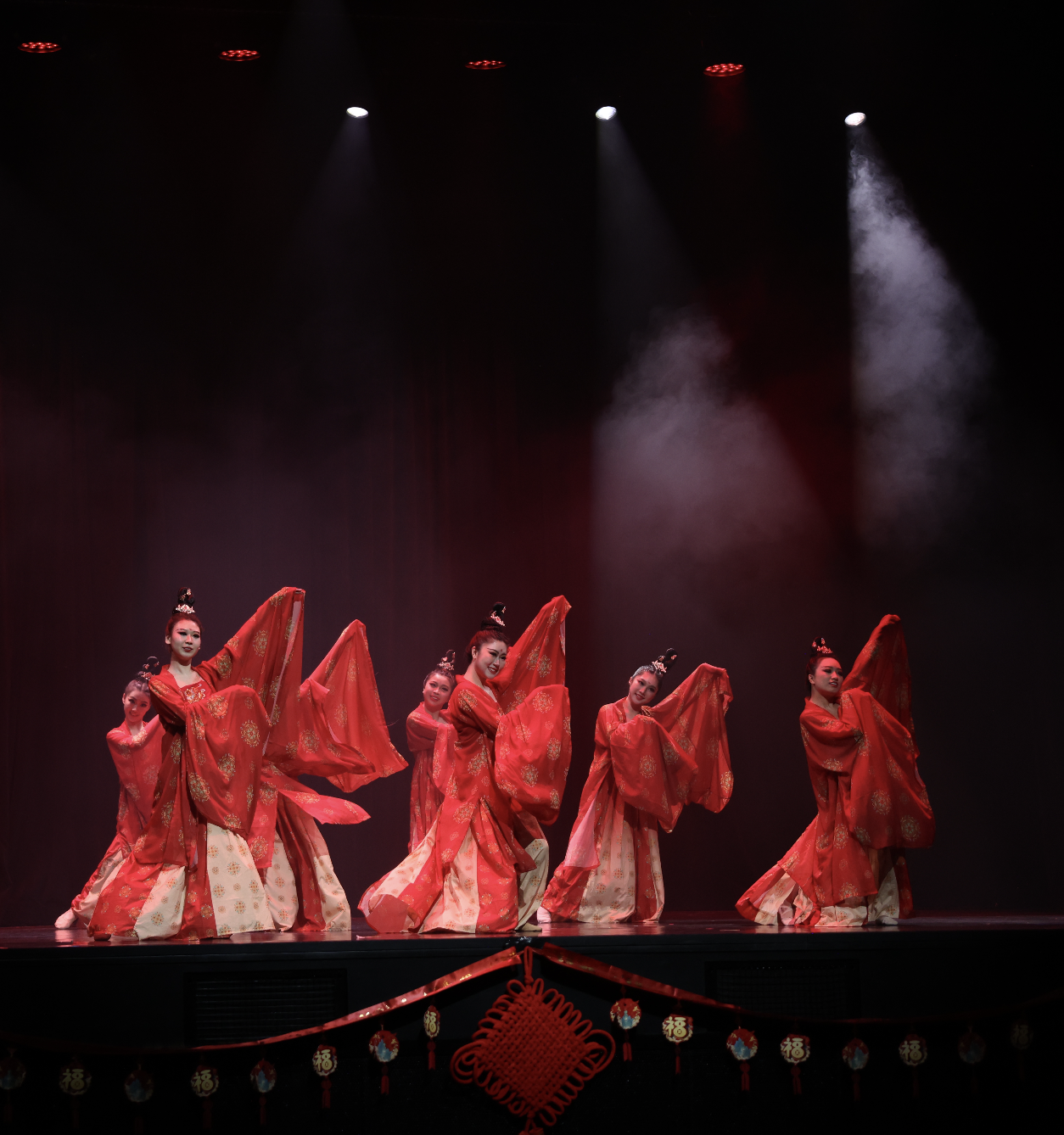 A group of female dancers dressed in traditional red and gold costumes performing on stage with dark background and stage lights.